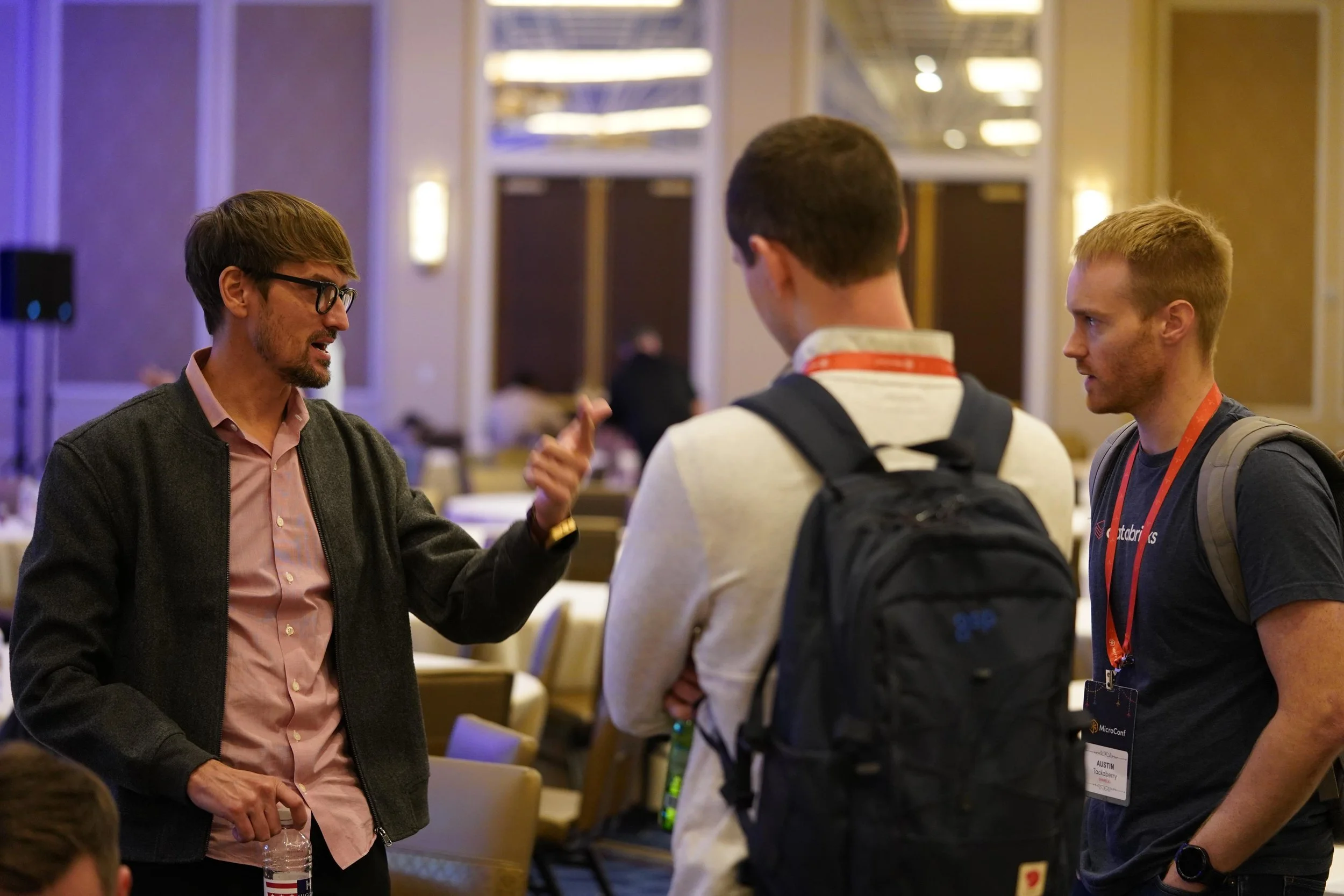 Three men engaged in a conversation in a conference or event setting. One man with glasses and a beard is speaking, while the other two men are listening, wearing name badges and backpacks.