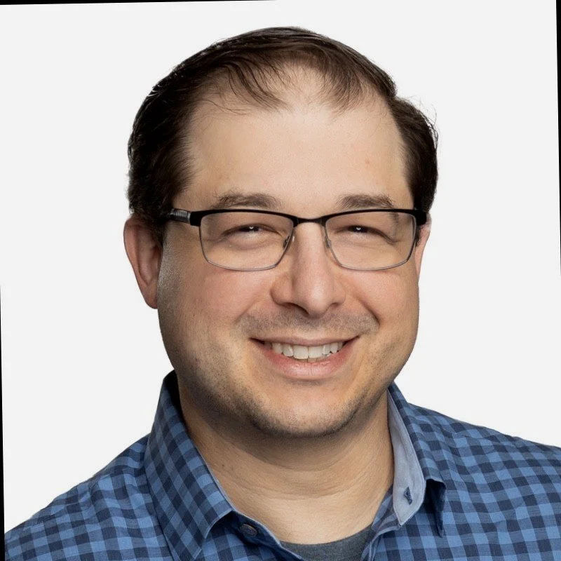 A smiling man with glasses wearing a blue checkered shirt against a plain white background.