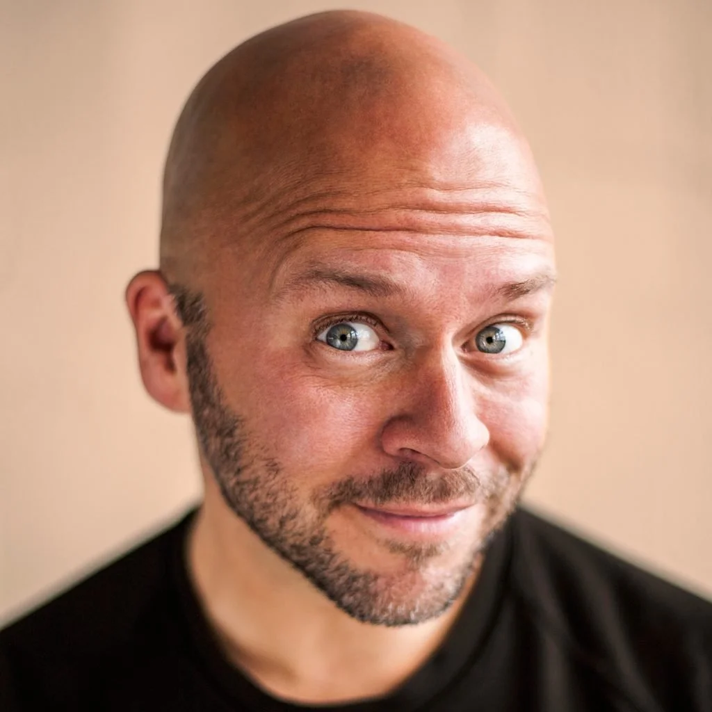 A bald man with blue eyes and a beard smiling at the camera, wearing a black shirt against a beige background.
