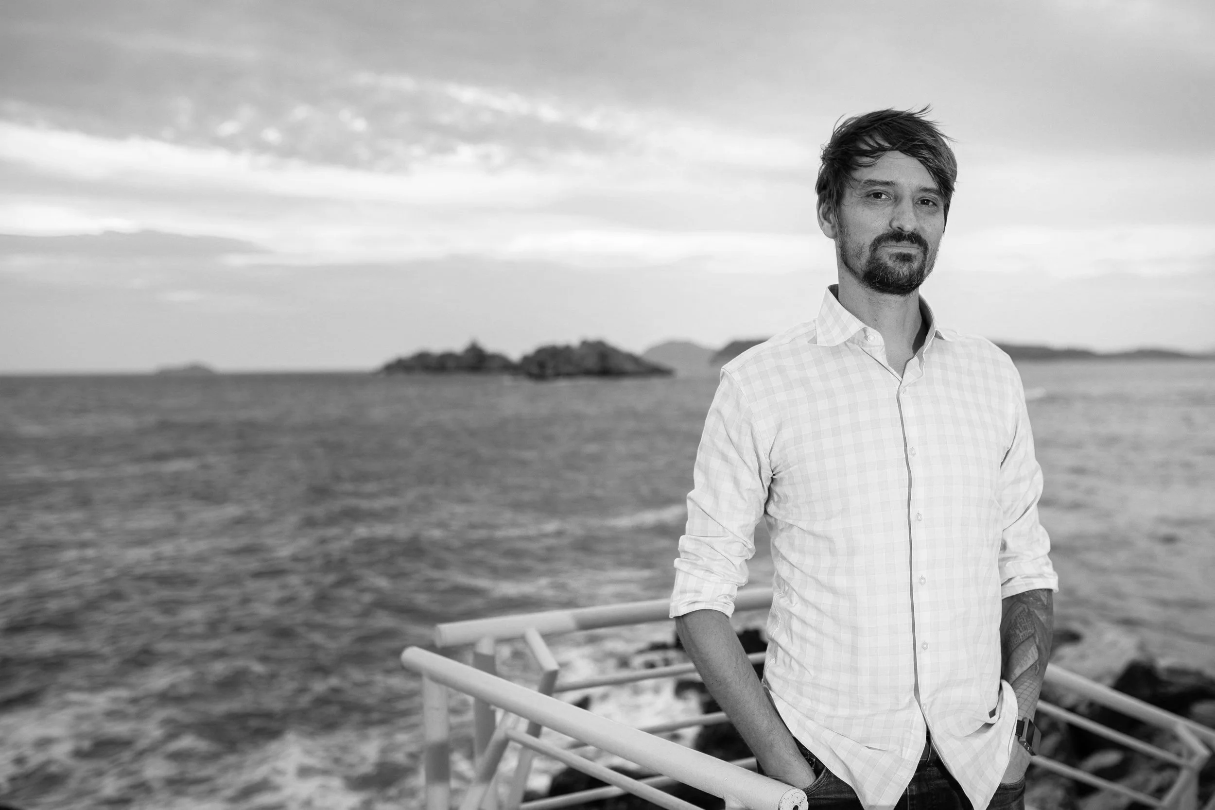 A black and white photo of a man standing on a beach with water and islands in the background. The man has a beard, shouldered-length hair, and is wearing a light checkered shirt with the sleeves rolled up.