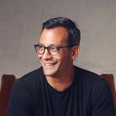 A smiling man with glasses wearing a blue checkered shirt against a plain white background.