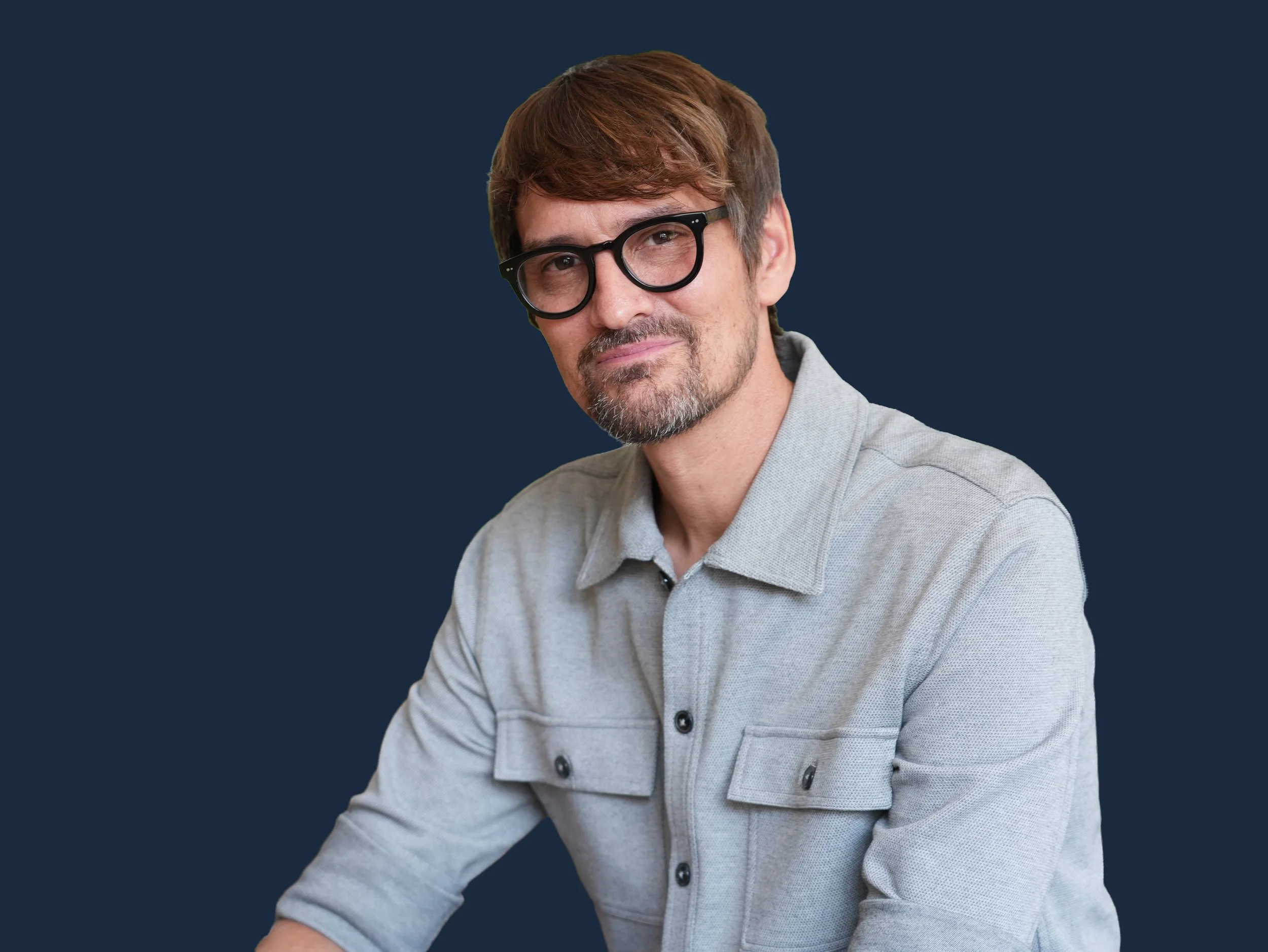 Portrait of a man with brown hair, glasses, and a beard, wearing a light gray shirt, against a dark blue background.