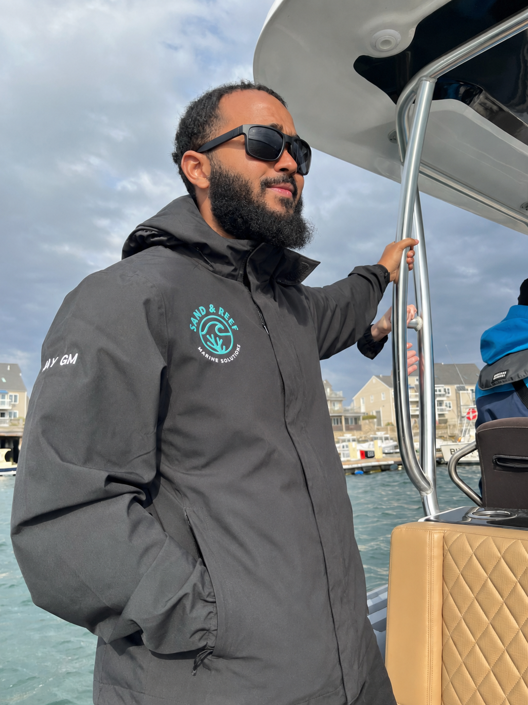 A man with a beard wearing sunglasses and a gray Sand & Reef Marine Solutions jacket, standing on a boat and holding onto a metal railing, with marina buildings in the background and a cloudy sky.
