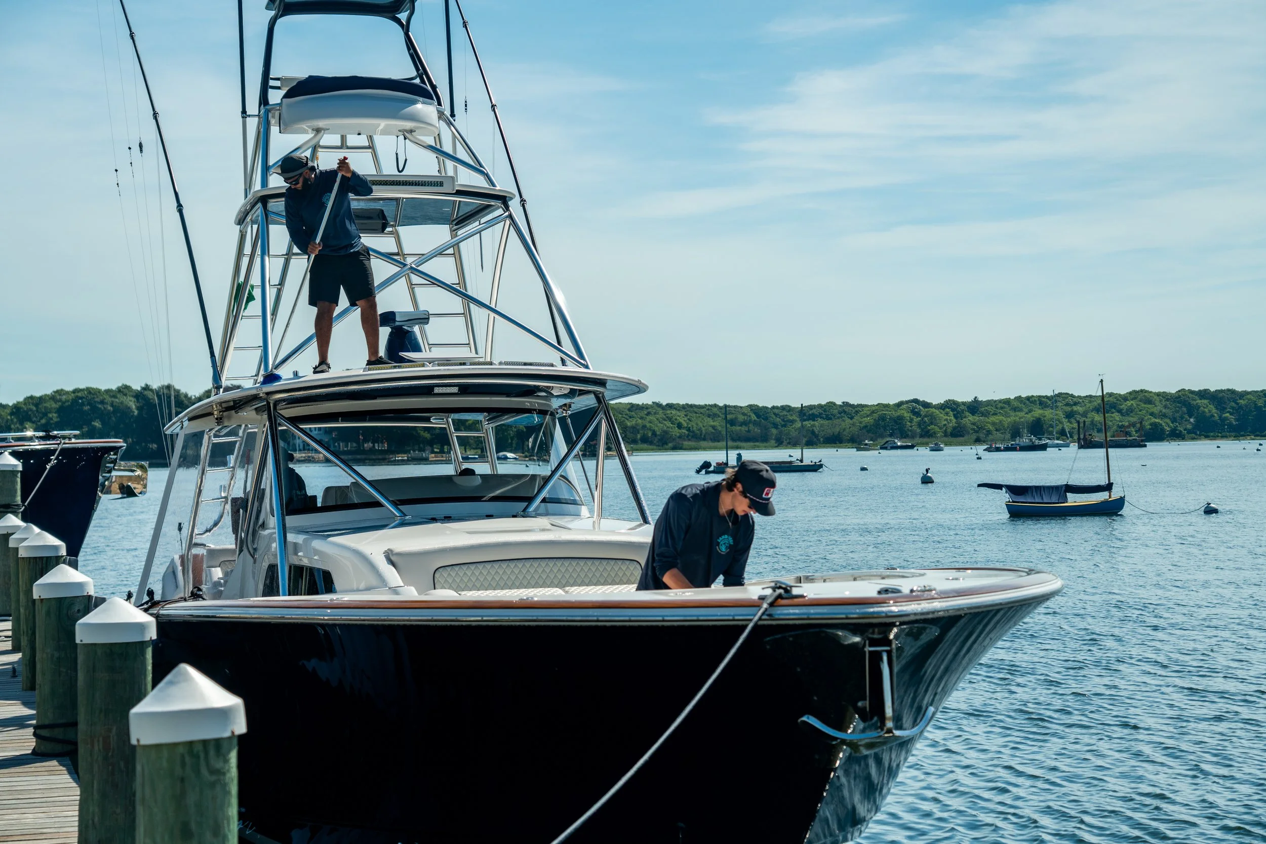 Two men working on a large yacht docked at a marina, with a calm water body, small boats, and a green treeline in the background under a partly cloudy sky.