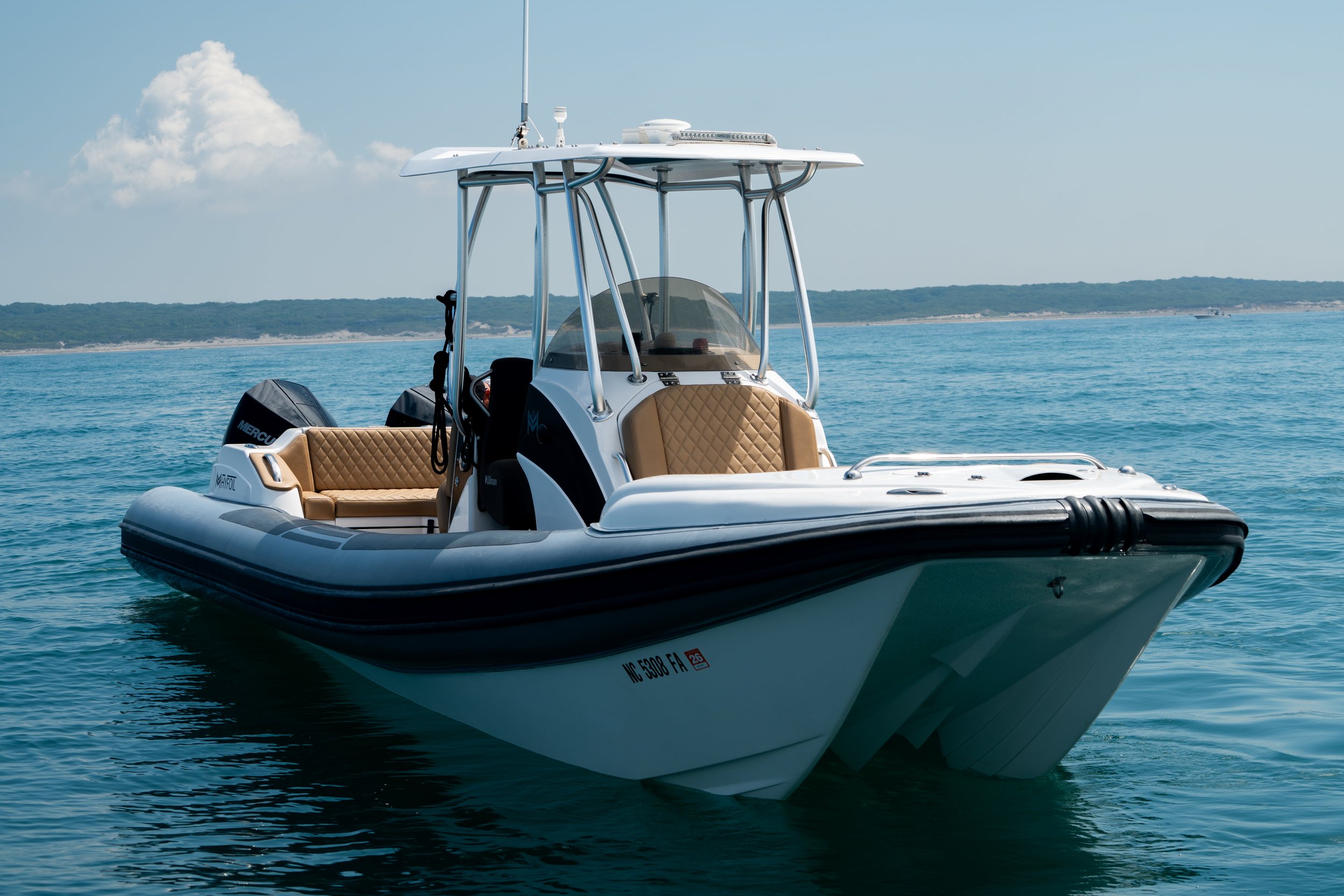 A white motorboat with beige quilted seats and a black Mercury outboard motor floats on calm blue water under a partly cloudy sky. In the background, a distant shoreline with green hills is visible.
