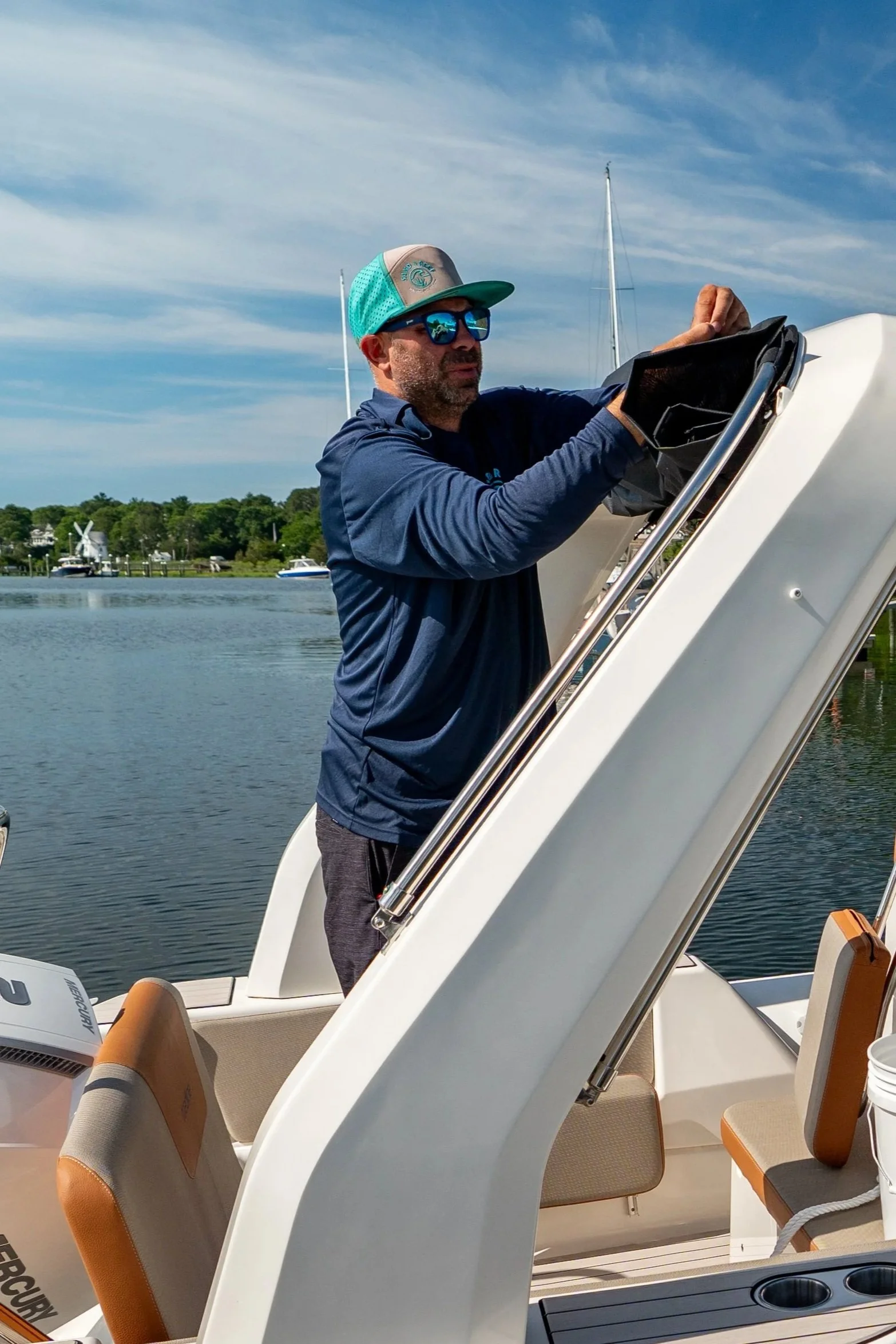 Man wearing sunglasses and a cap, working on a boat with water, sailboats, and trees in the background.