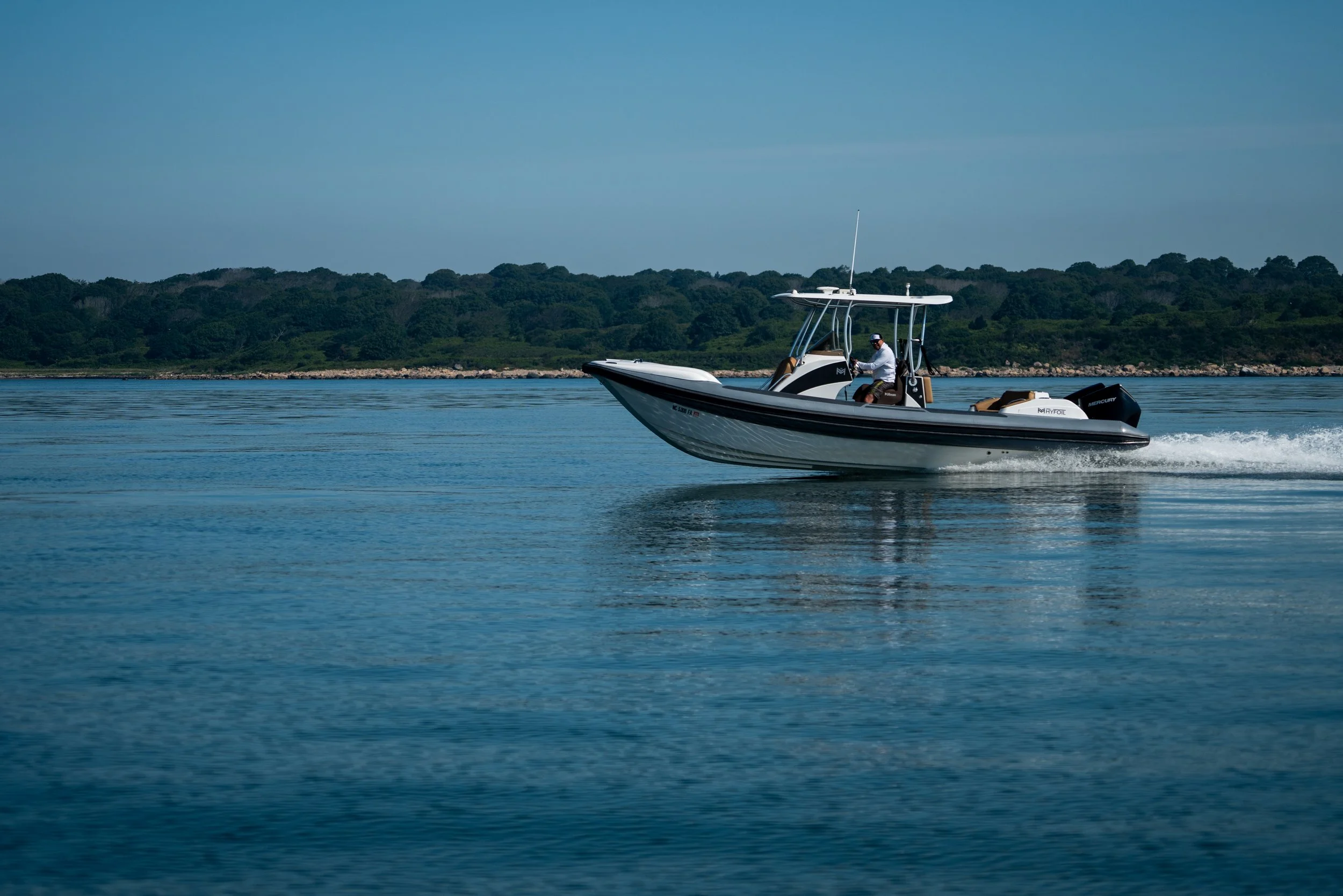 A small motorboat moving across a body of water, with a green tree-covered shoreline in the background and a clear blue sky overhead.