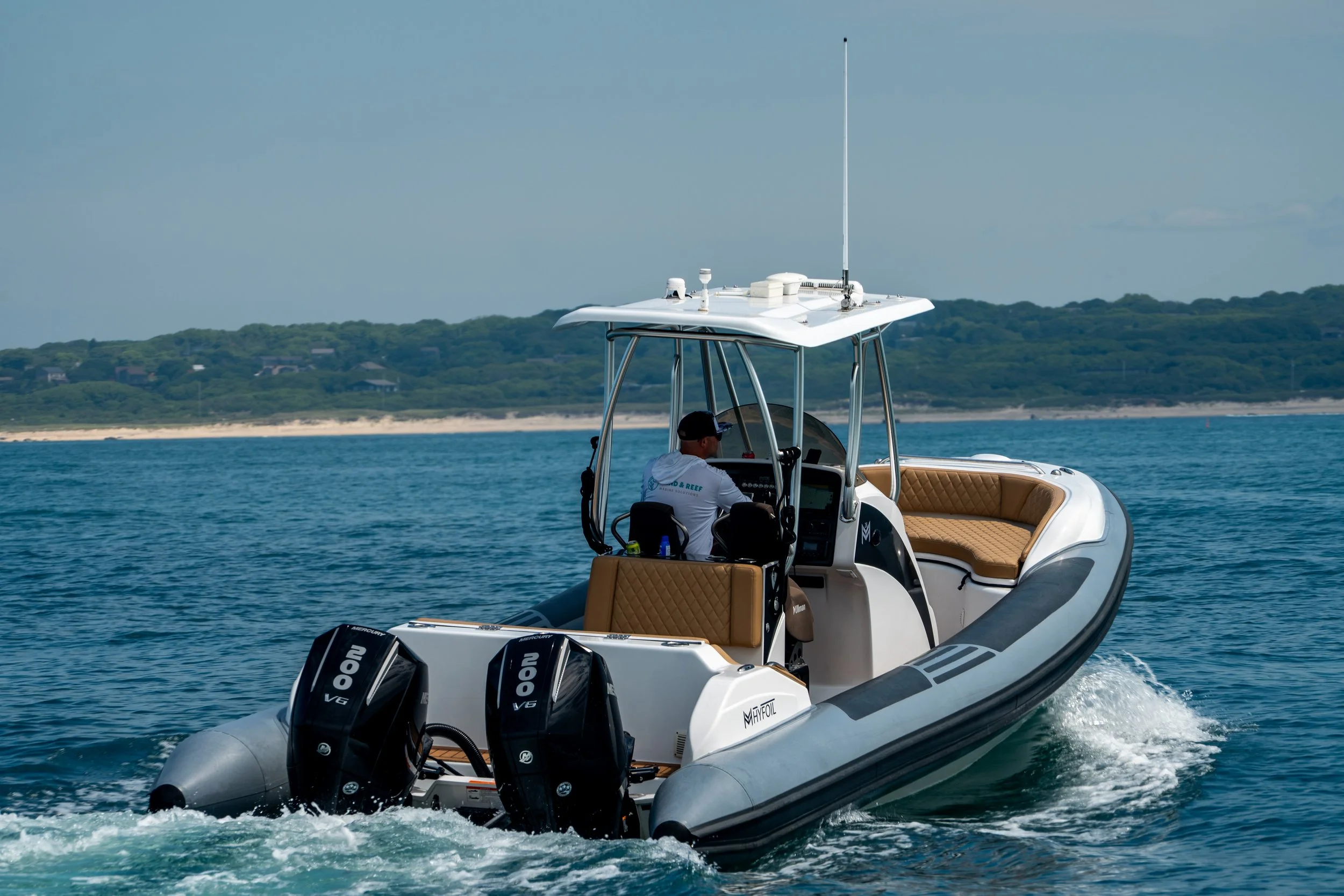 A white and gray motorboat with a covered dashboard navigating on blue water near a coastline with green hills and houses.