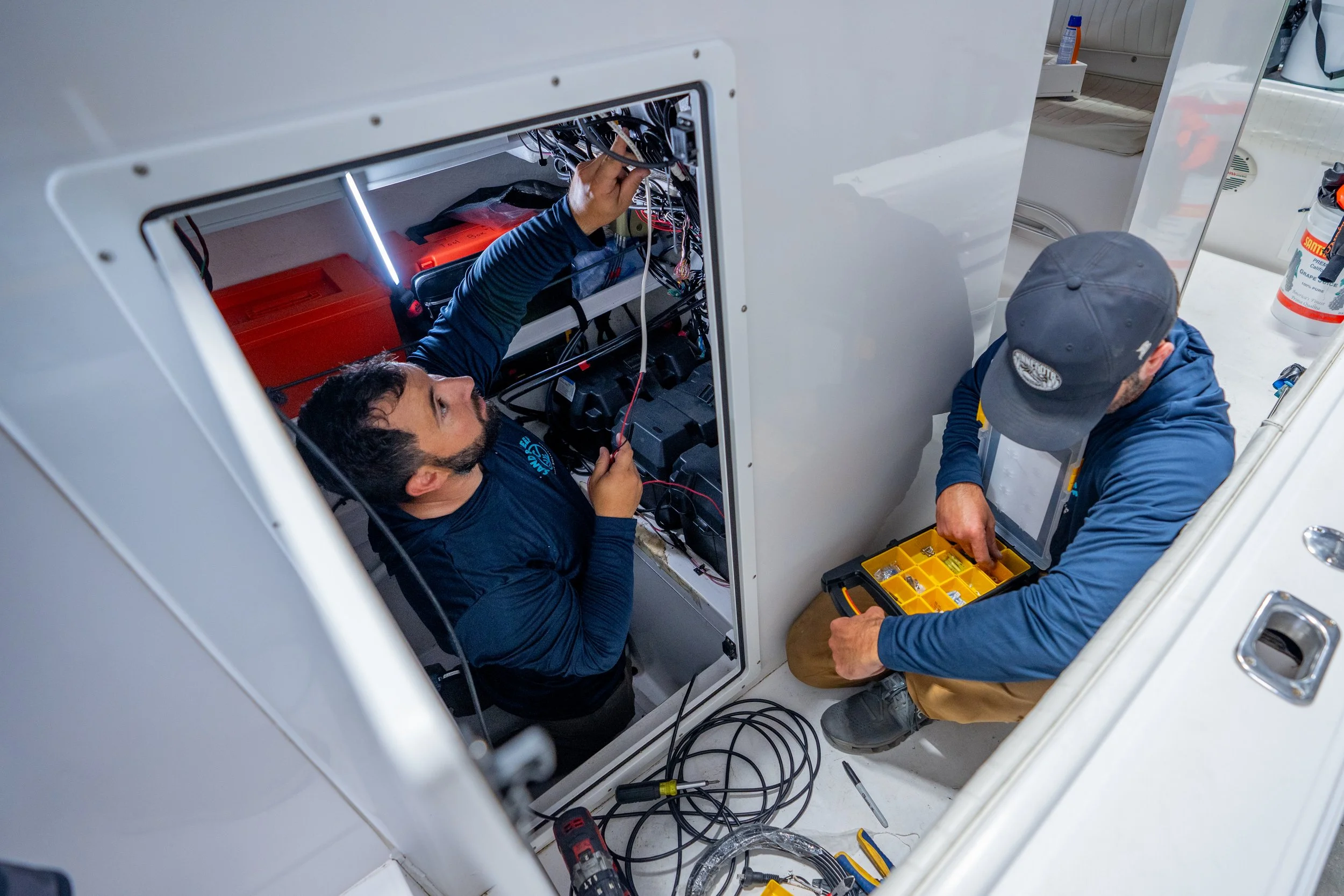 Two technicians working on electrical wiring inside a laundry machine, with tools and a yellow parts organizer nearby.