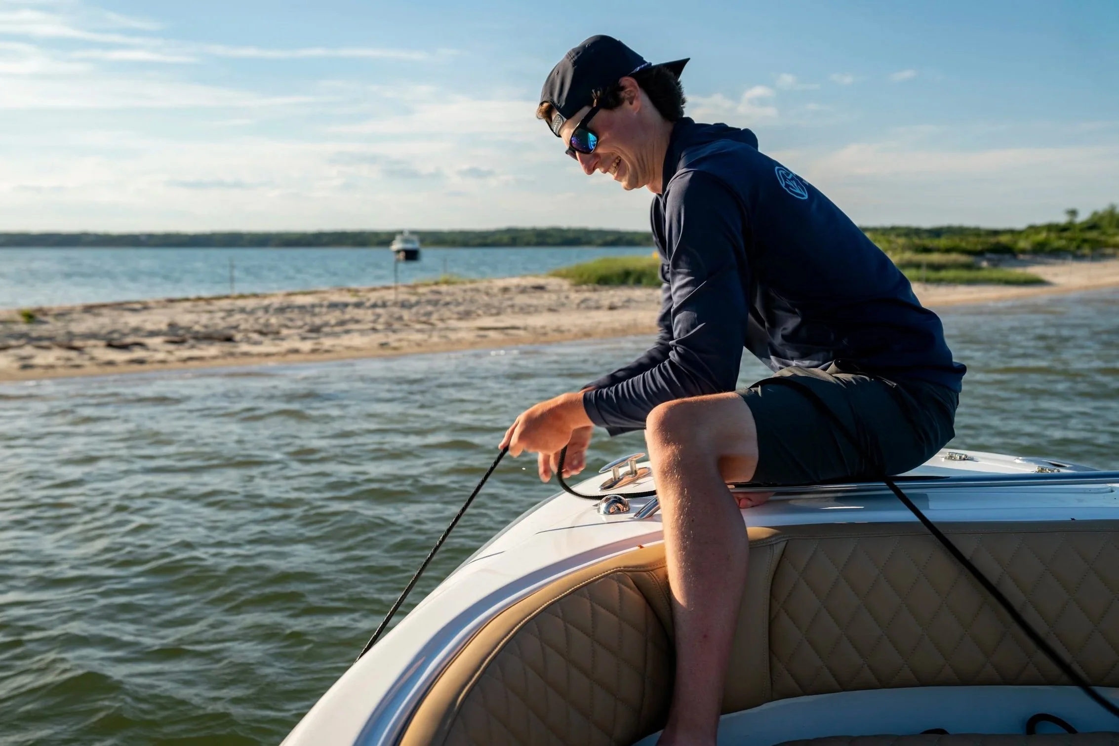 A man smiling and wearing sunglasses, a black cap, and a dark jacket, sitting on a boat near a shoreline with a boat in the water and a clear sky.