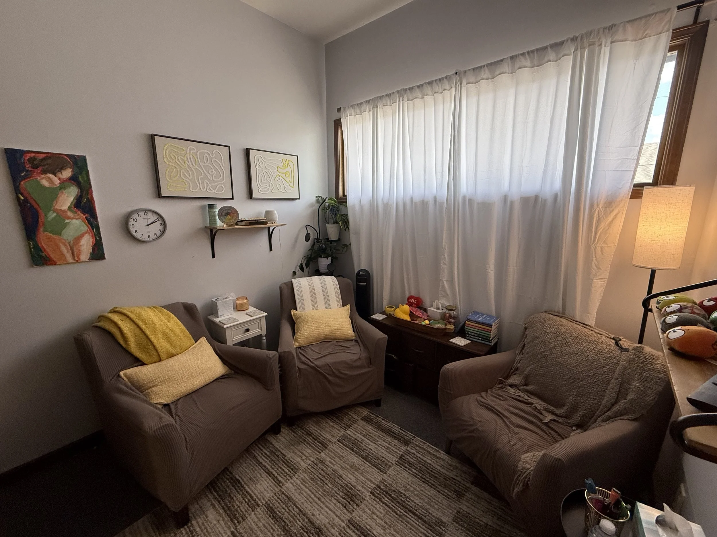 A cozy living room with two brown sofas and yellow throw pillows, a white side table, wall art, a wall clock, and a large window with white curtains. There's a floor lamp, a plant, toys, and a bookshelf with books.