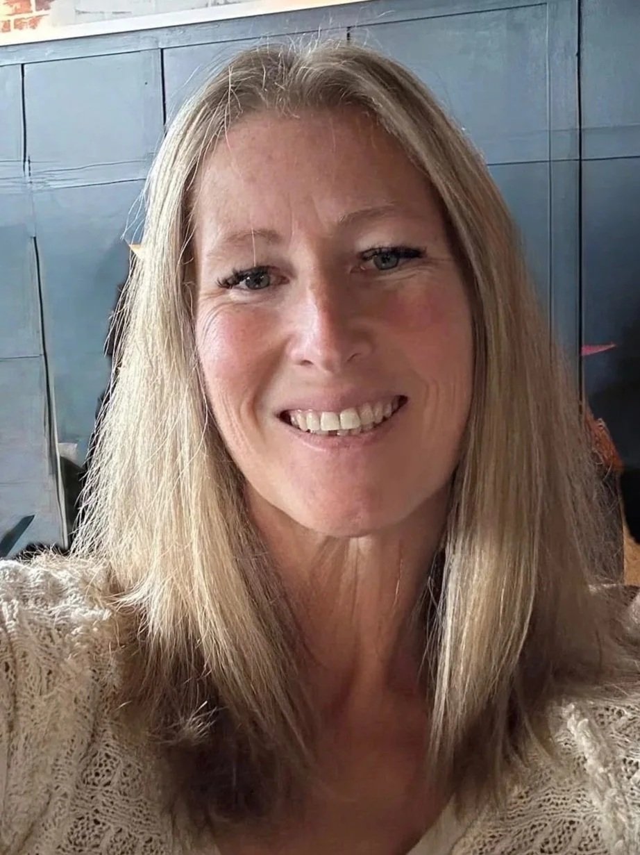 A smiling woman with long blonde hair wearing a lace top in an indoor setting.