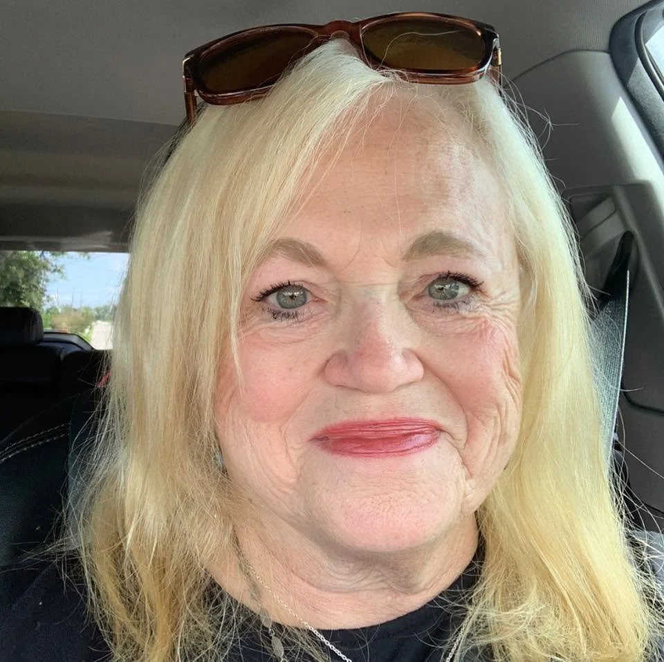 A smiling older woman with blonde hair and sunglasses on her head, sitting in a car.