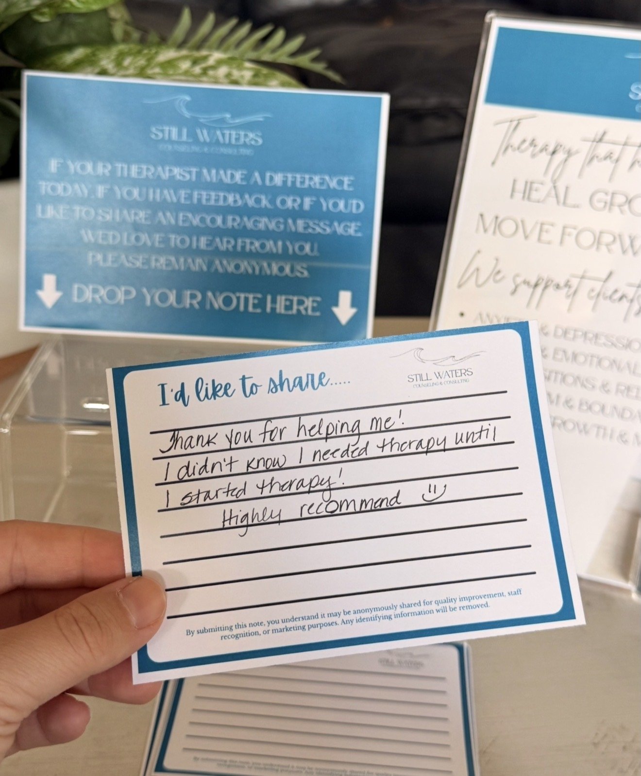 Close-up of a blue and white note card with handwritten message expressing gratitude for therapy. In the background, there are other similar notice boards with motivational and inspirational messages about healing, moving forward, and gratitude. The setting appears to be a mental health or support center.