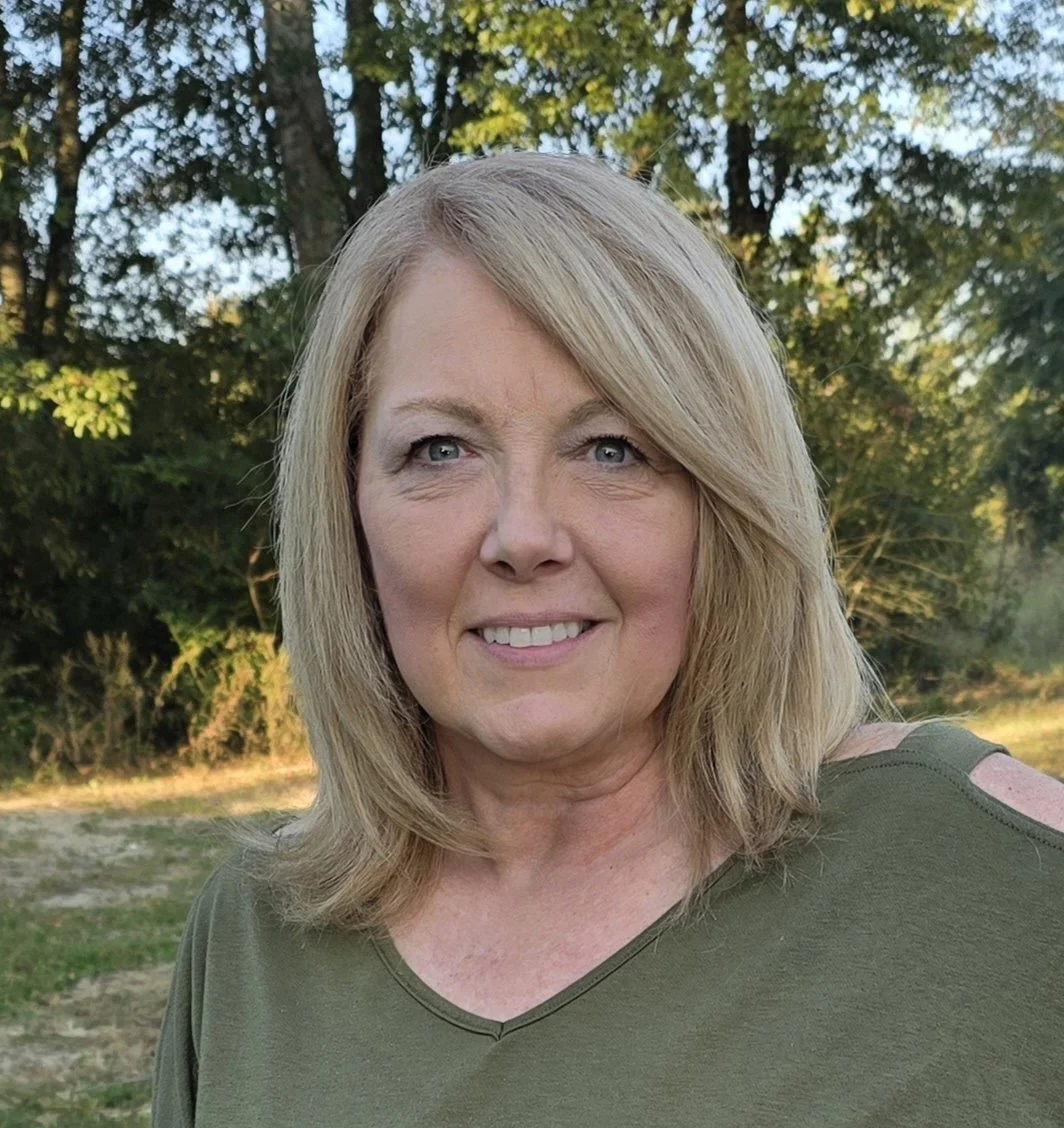 A woman with blonde hair and blue eyes smiling outdoors with trees in the background.