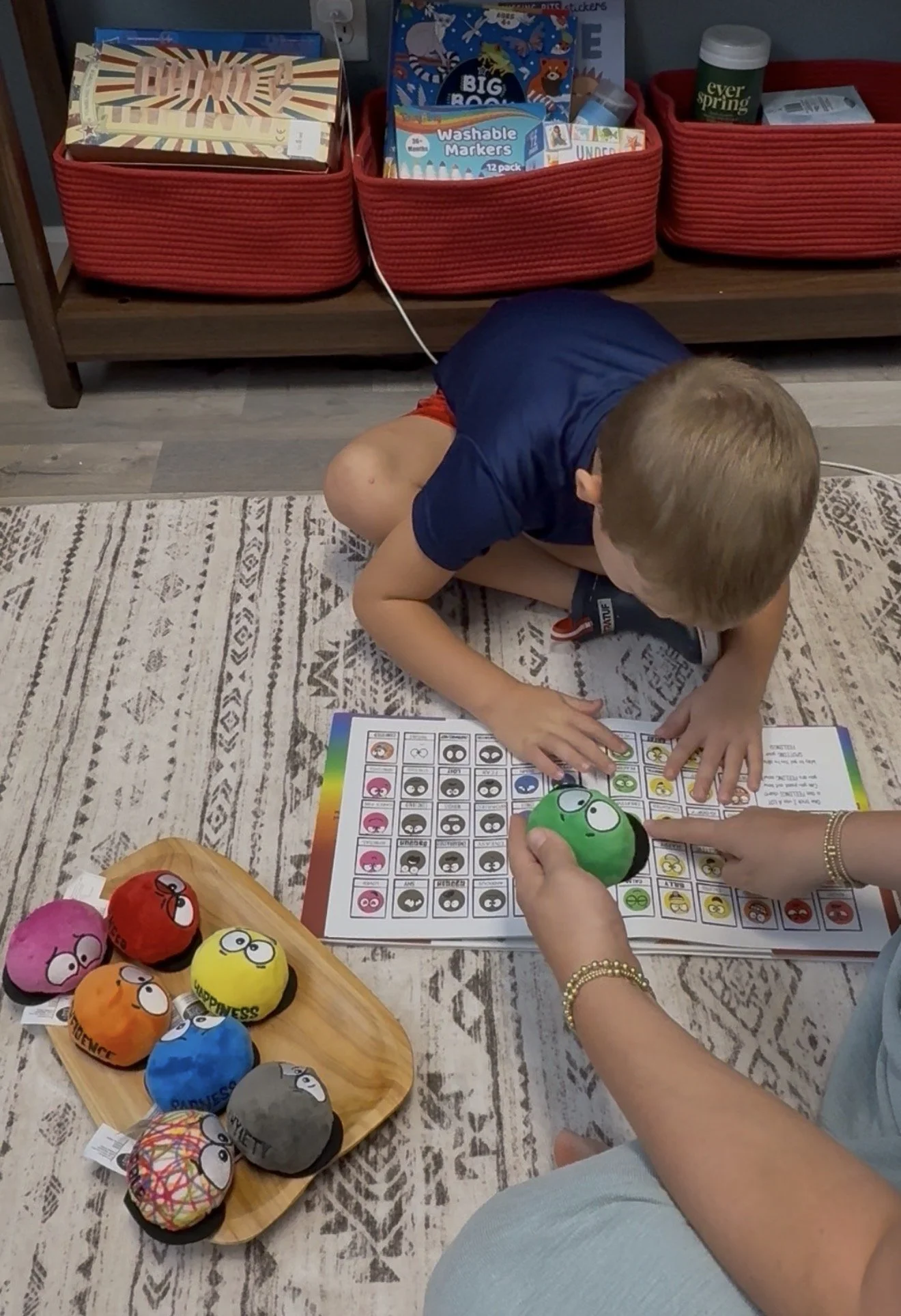 Child sitting on the floor playing a bingo game with colorful plush balls labeled with words like 'Happiness' and 'Adventure' on a wooden tray nearby. The game involves matching colored and numbered balls to a bingo chart, with an adult pointing at the chart. The setting includes red storage baskets filled with books and other items on a shelf, and a patterned rug on the floor.