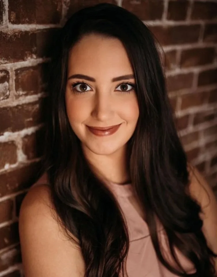 A young woman with long dark hair and light skin smiling at the camera, standing against a brick wall.
