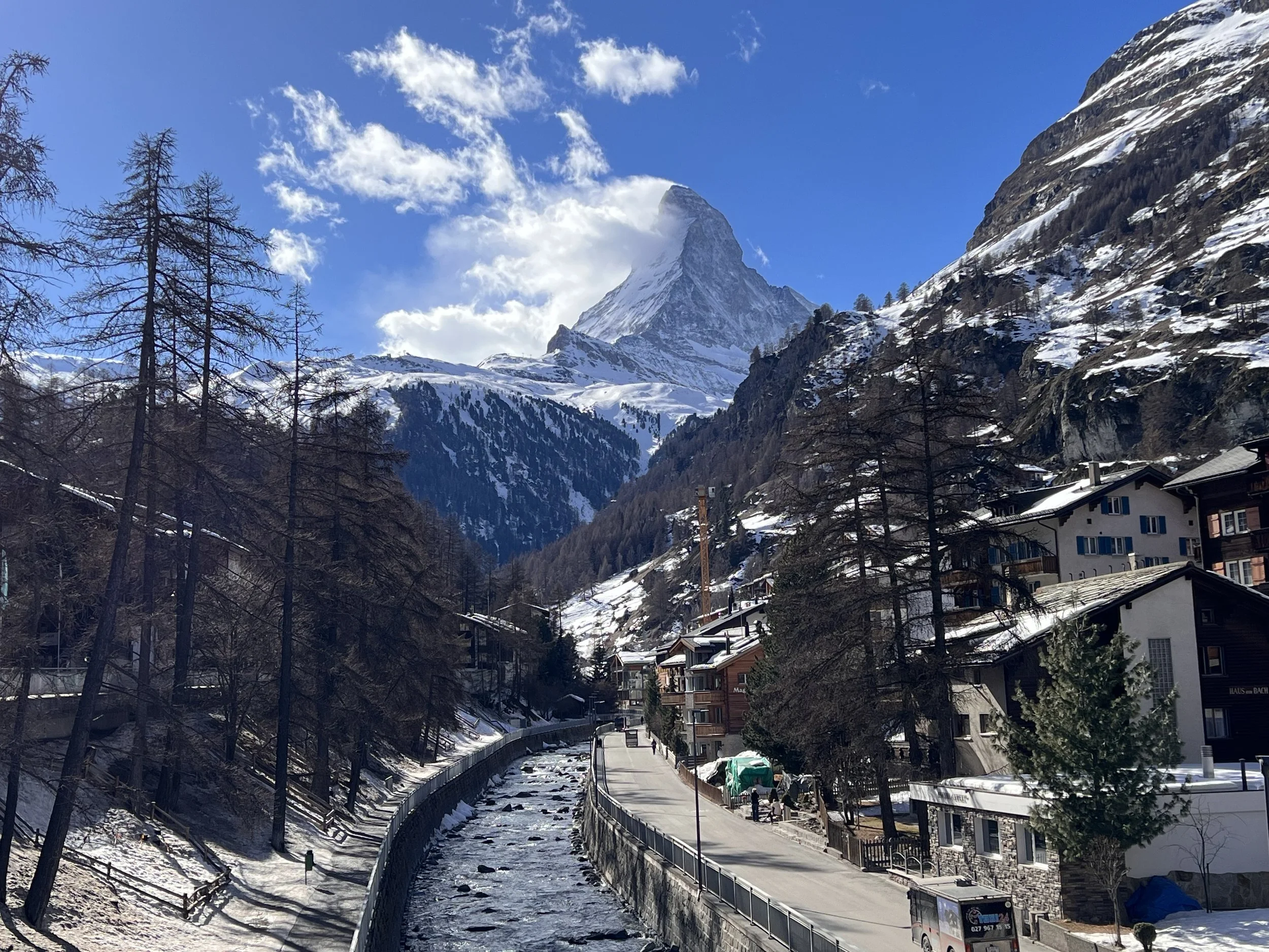 View of the Matterhorn rising above Zermatt, Switzerland, framed by pine trees, chalet rooftops, and the Vispa river running through the village below a partly cloudy blue sky.