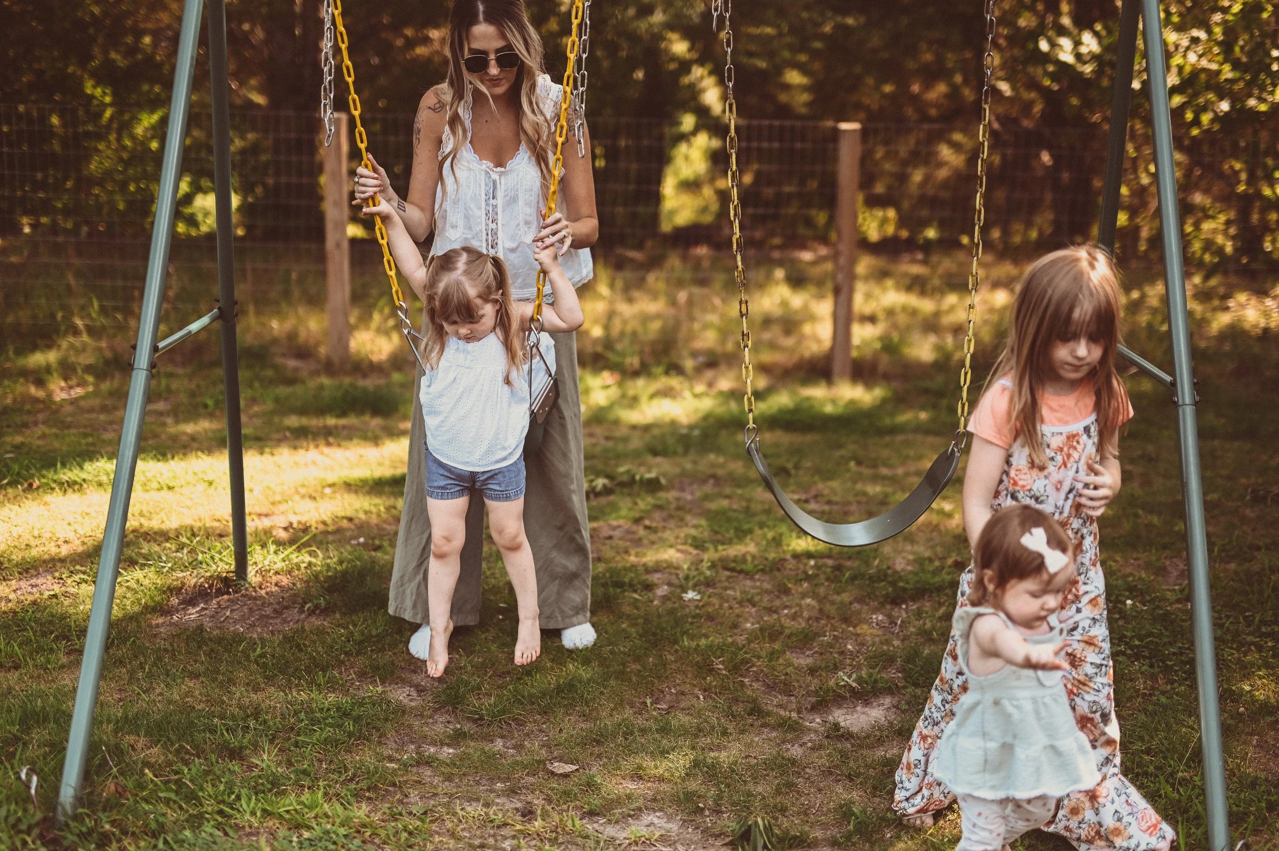 A woman with long hair, sunglasses, and a white sleeveless top is standing behind a girl on a swing set, holding the swing chains. The girl with curly hair wearing a white top and blue shorts appears to be about to swing or step off the swing. Anothe