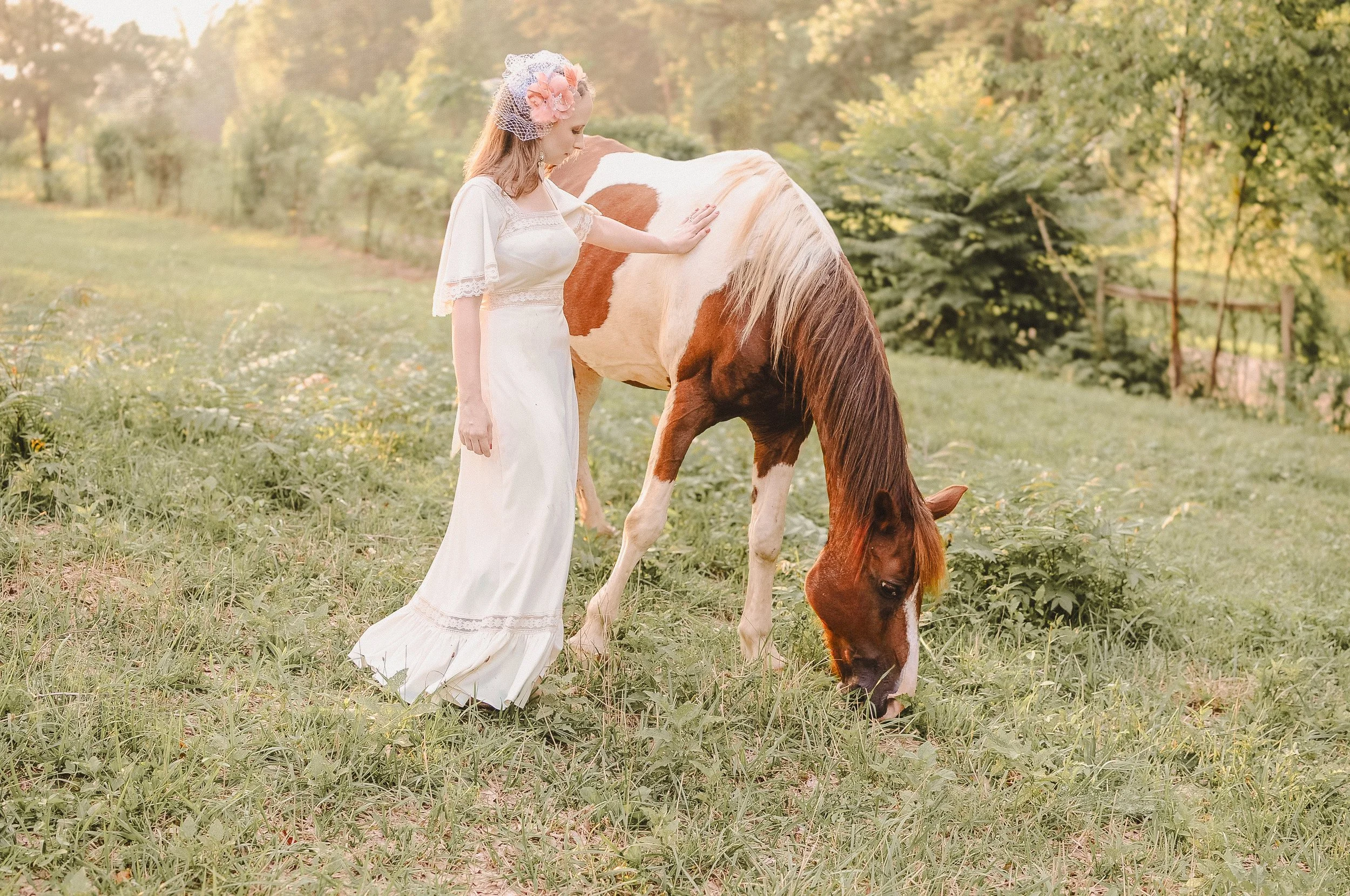 A woman in a white dress gently touching a brown and white horse in a grassy field with trees in the background.