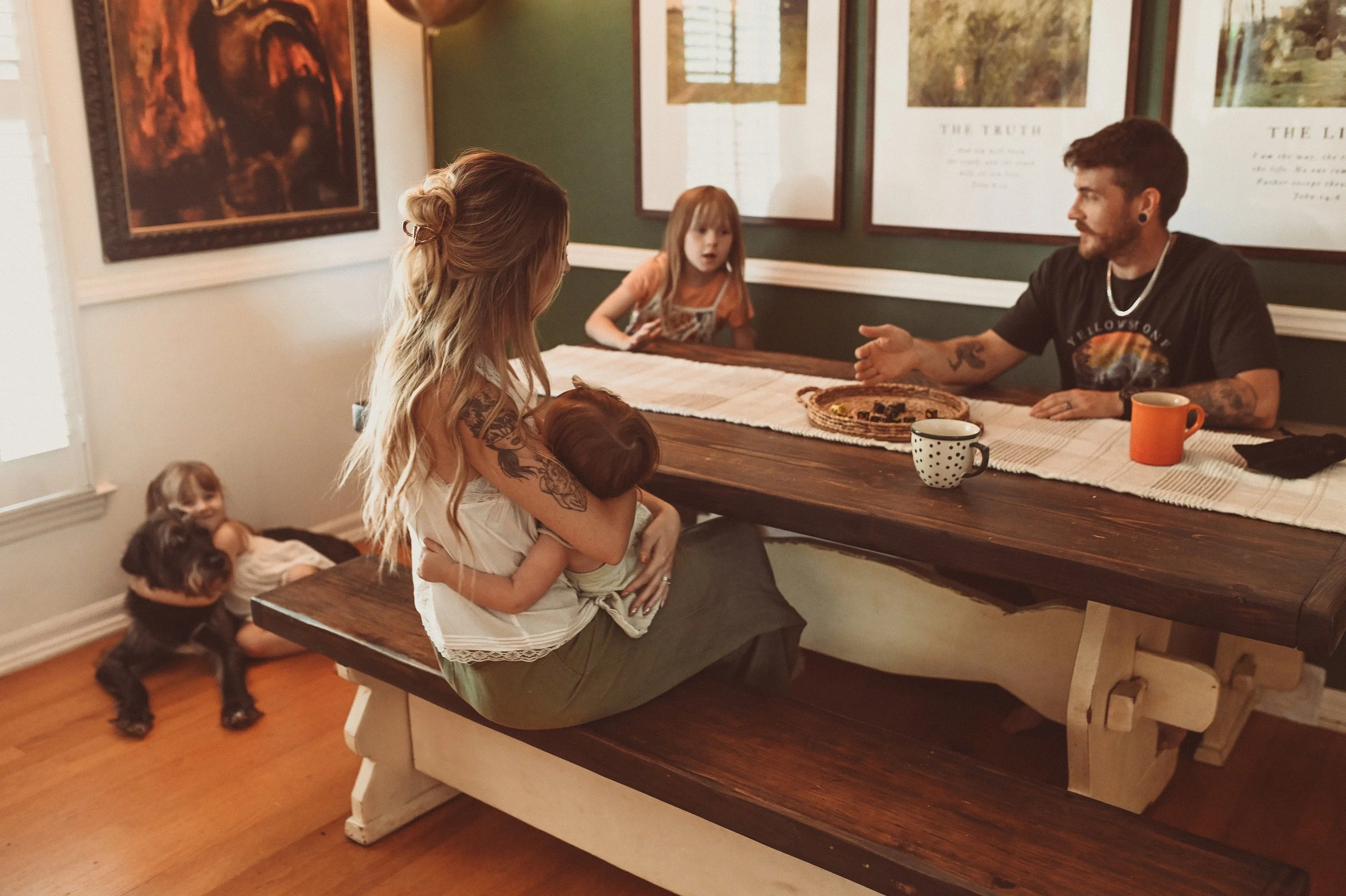 A woman with long, wavy blonde hair, tattoos on her arm, holding a child, sits at a dining table with three children nearby. One child is seated at the table with a surprised expression, and two children are on the floor near the table, one hugging a