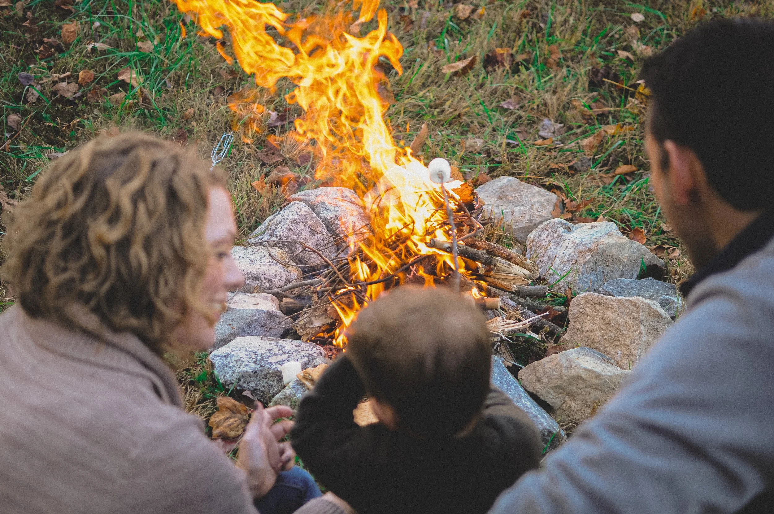People gathering around a small campfire outdoors on a rocky surface surrounded by fallen leaves.
