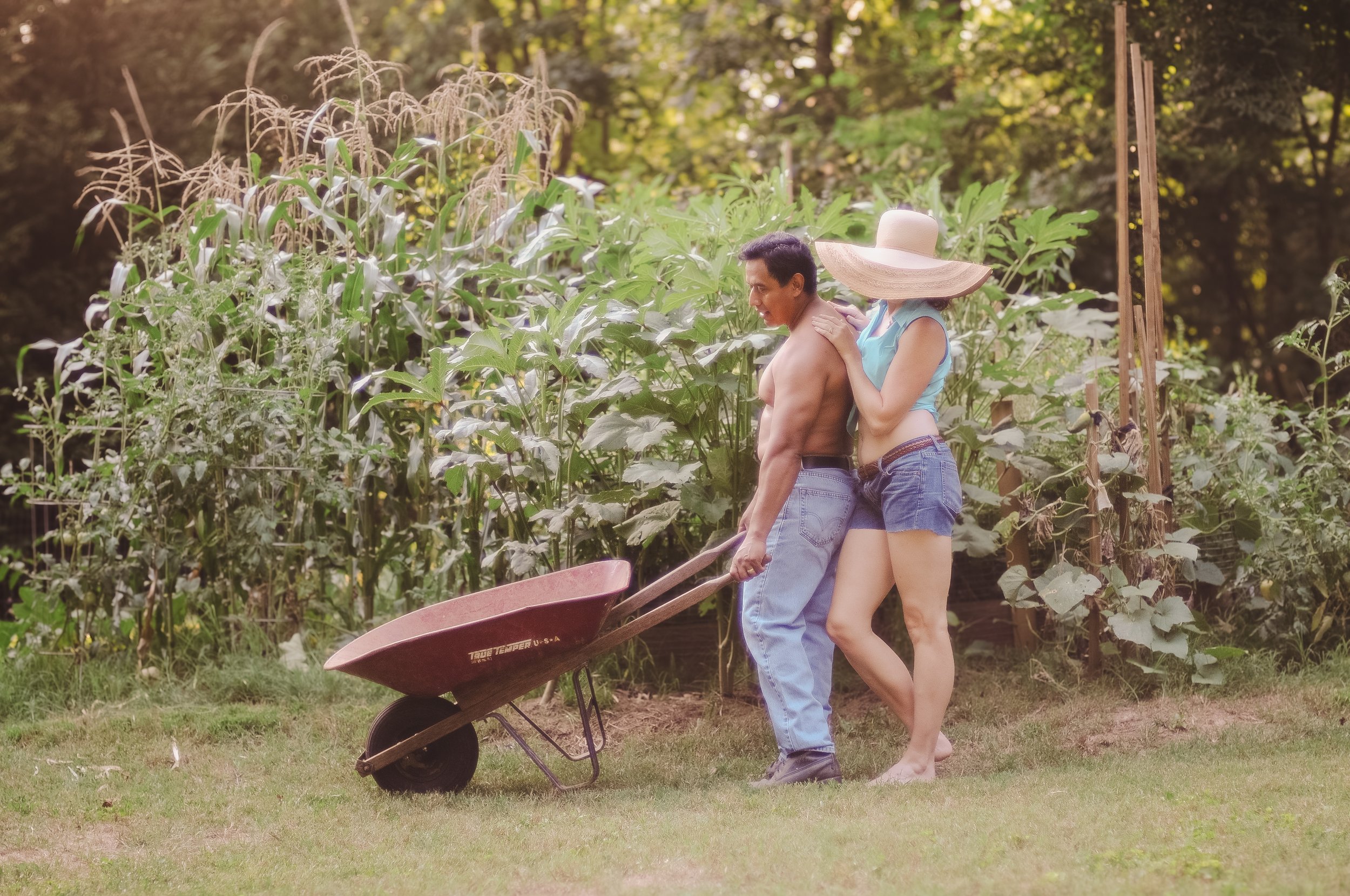 A man and woman gardening together in a backyard, with the woman leaning on the man’s shoulder while they push a wheelbarrow filled with plants.