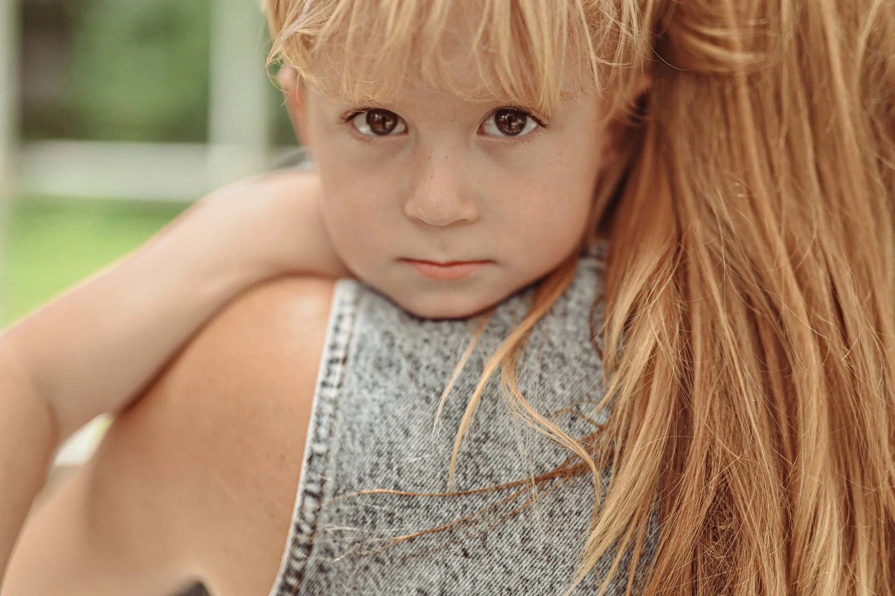 A young child with red hair and brown eyes watching the camera, resting their chin on someone with long hair, outdoors with a blurred green background.