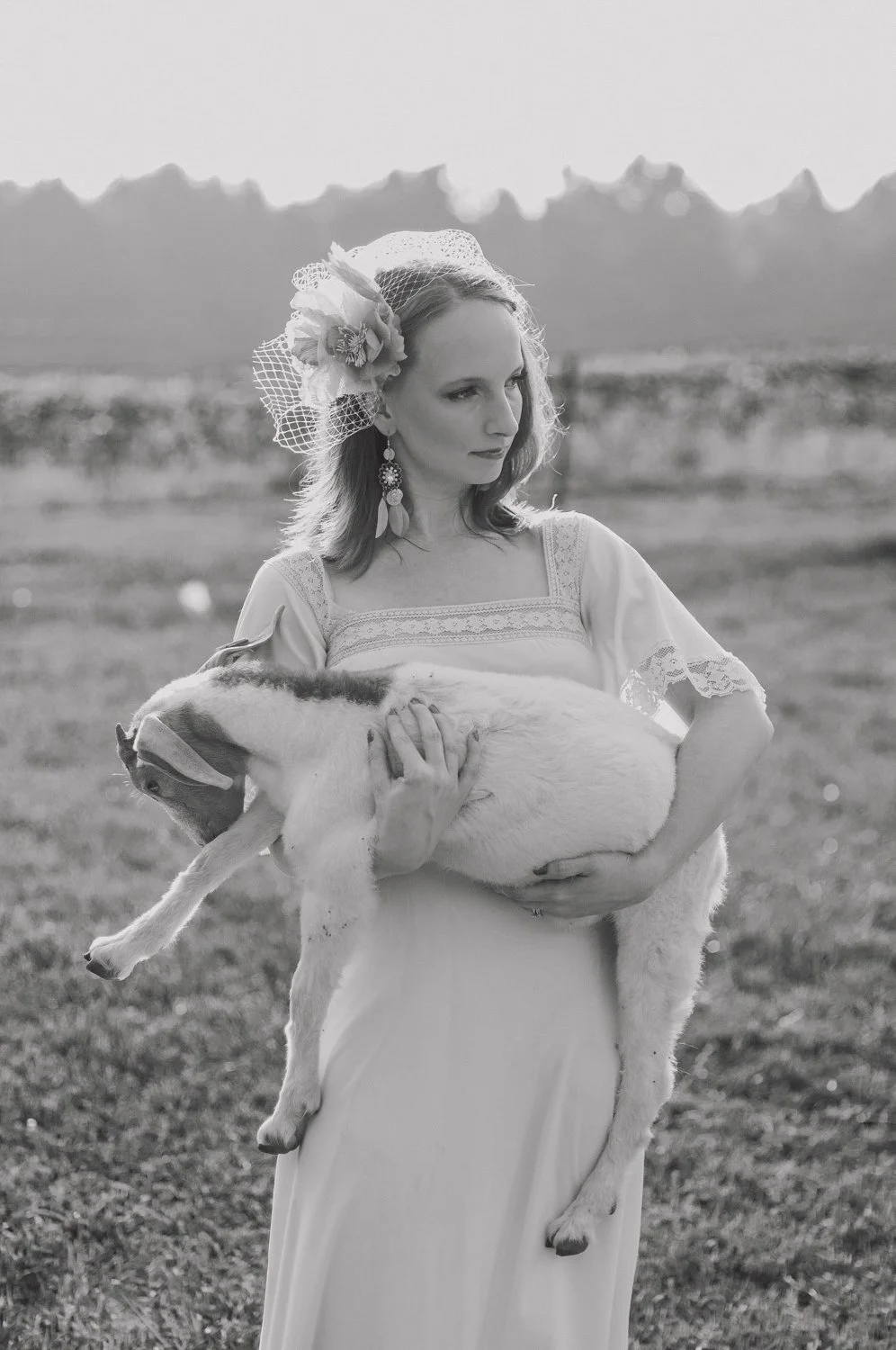 A woman holding a puppy outdoors in a field, wearing a dress and a floral hat with netting, in black and white.