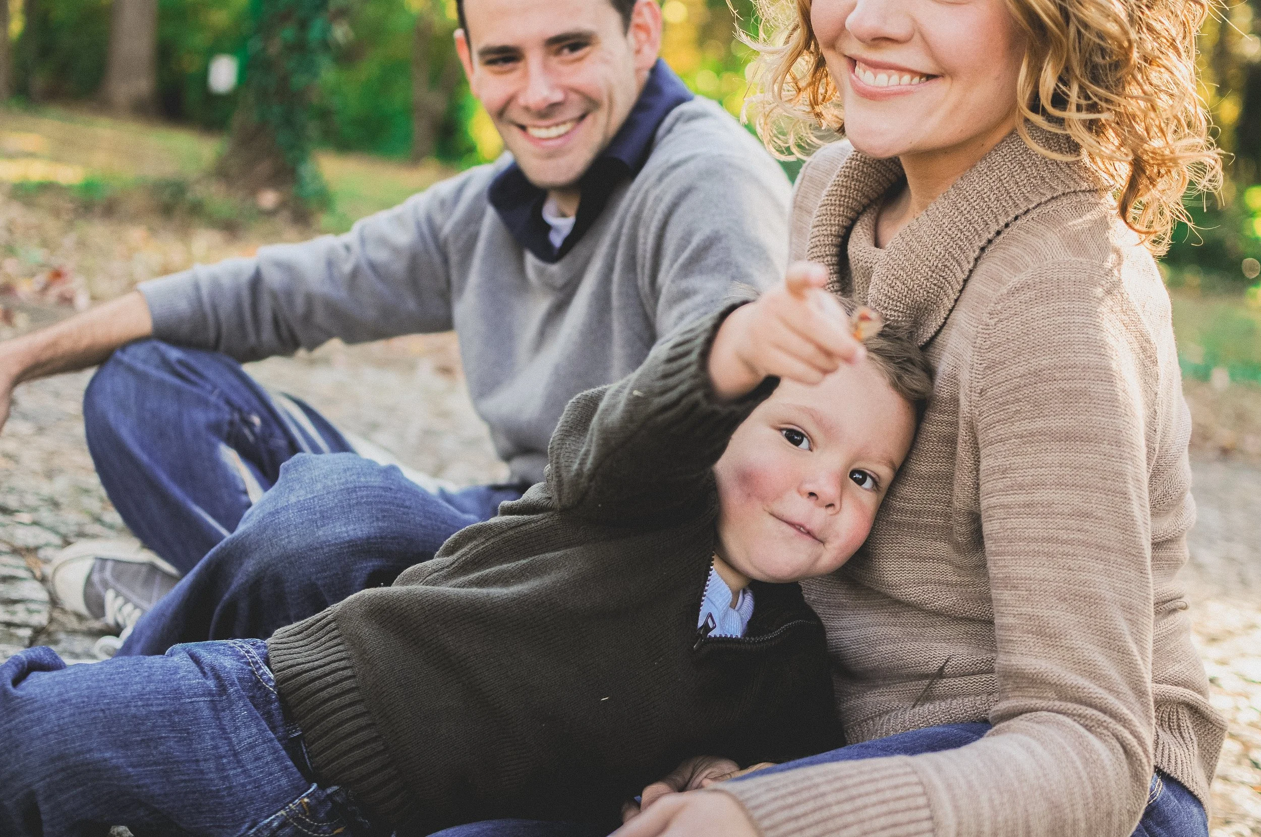 A smiling family with a man, a woman, and a young boy, sitting outdoors on a sunny day with trees and green foliage in the background.