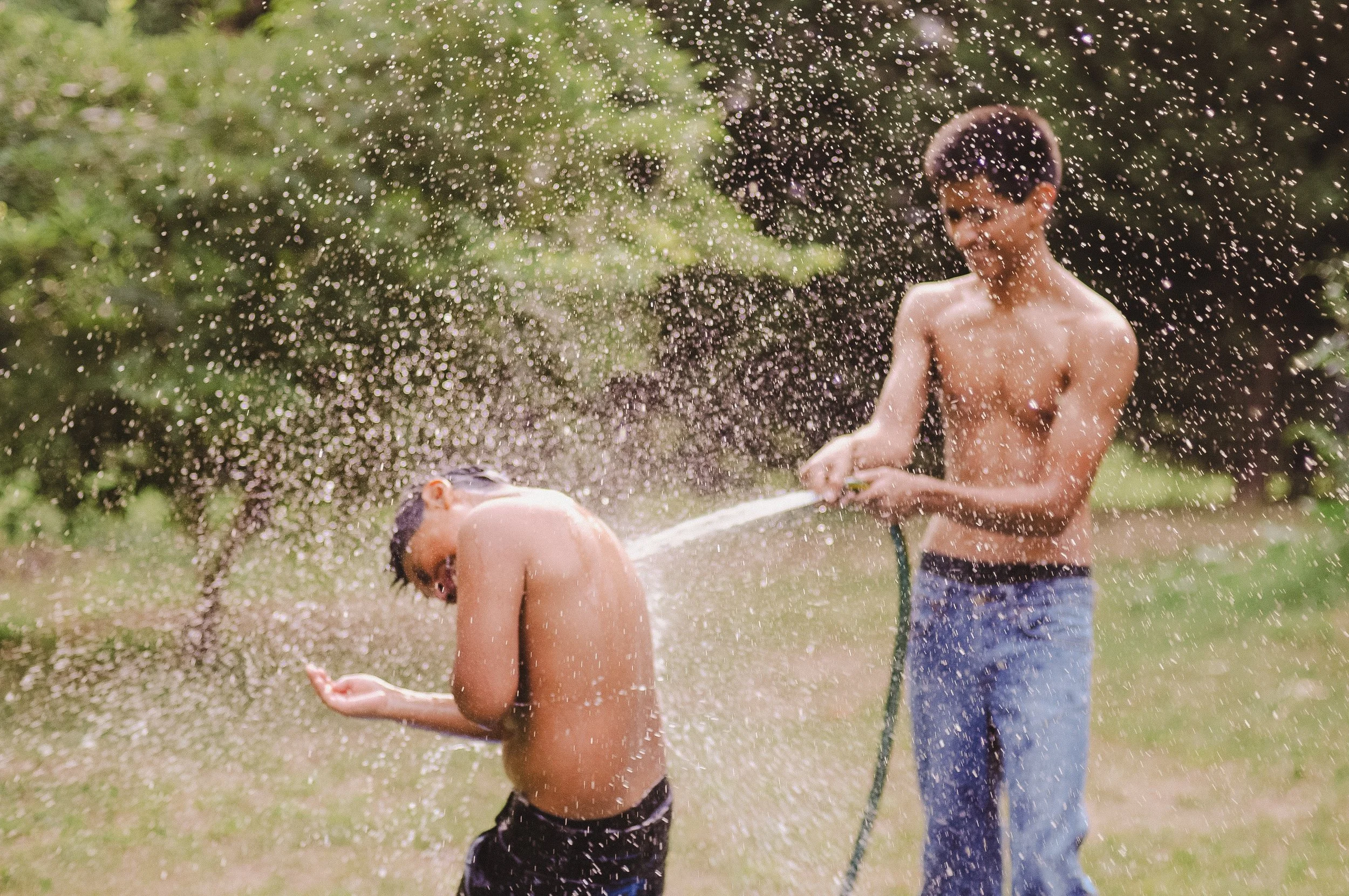 Two boys playing with water outdoors, one boy is shirtless leaning forward with water splashing on him, and the other boy is standing, holding a water hose, both surrounded by greenery.