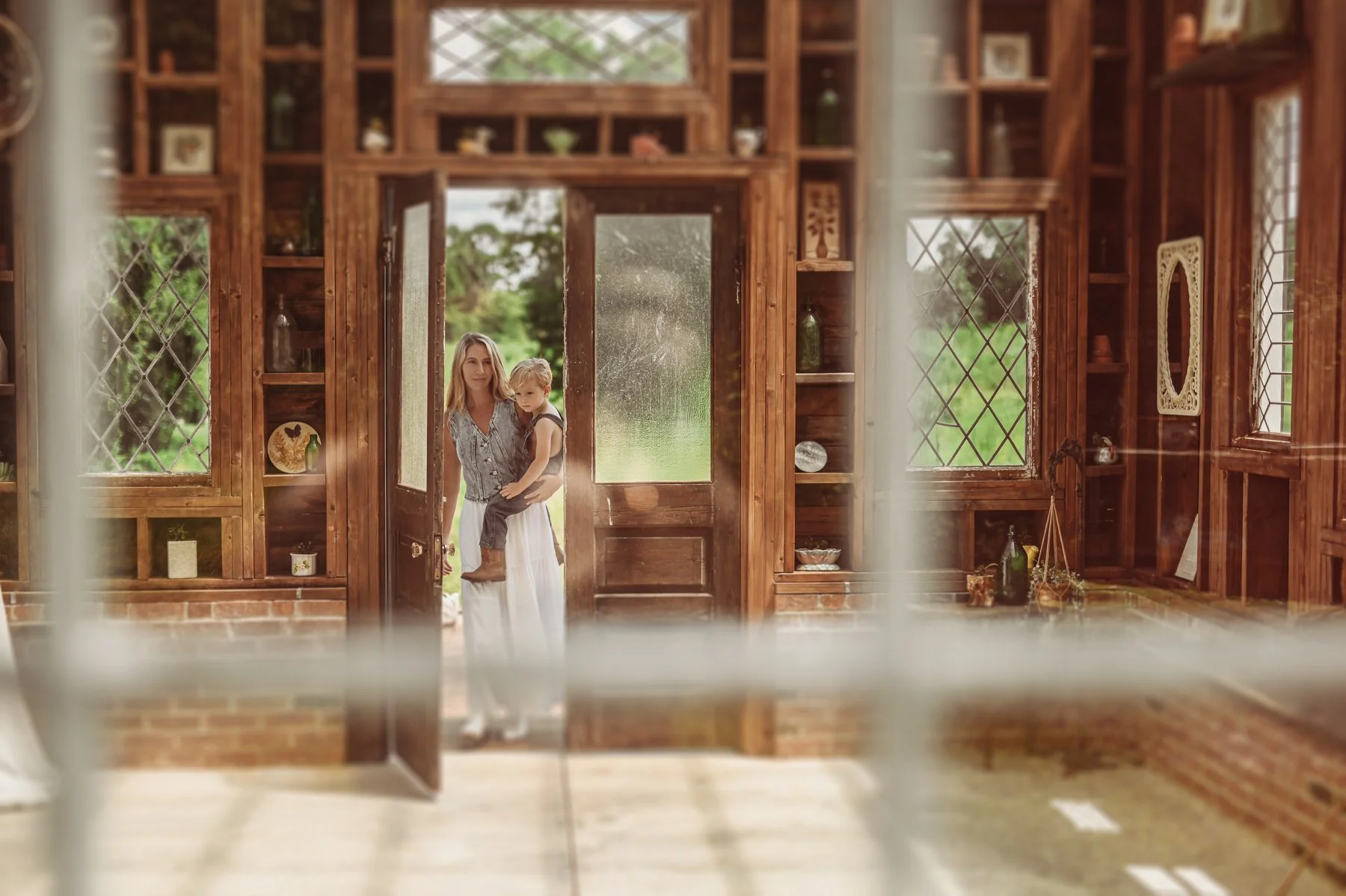 A woman is standing at the open door of a wooden, glass-paneled house, holding a young child in her arms. The scene is viewed through a blurred window screen with a green outdoor background visible.