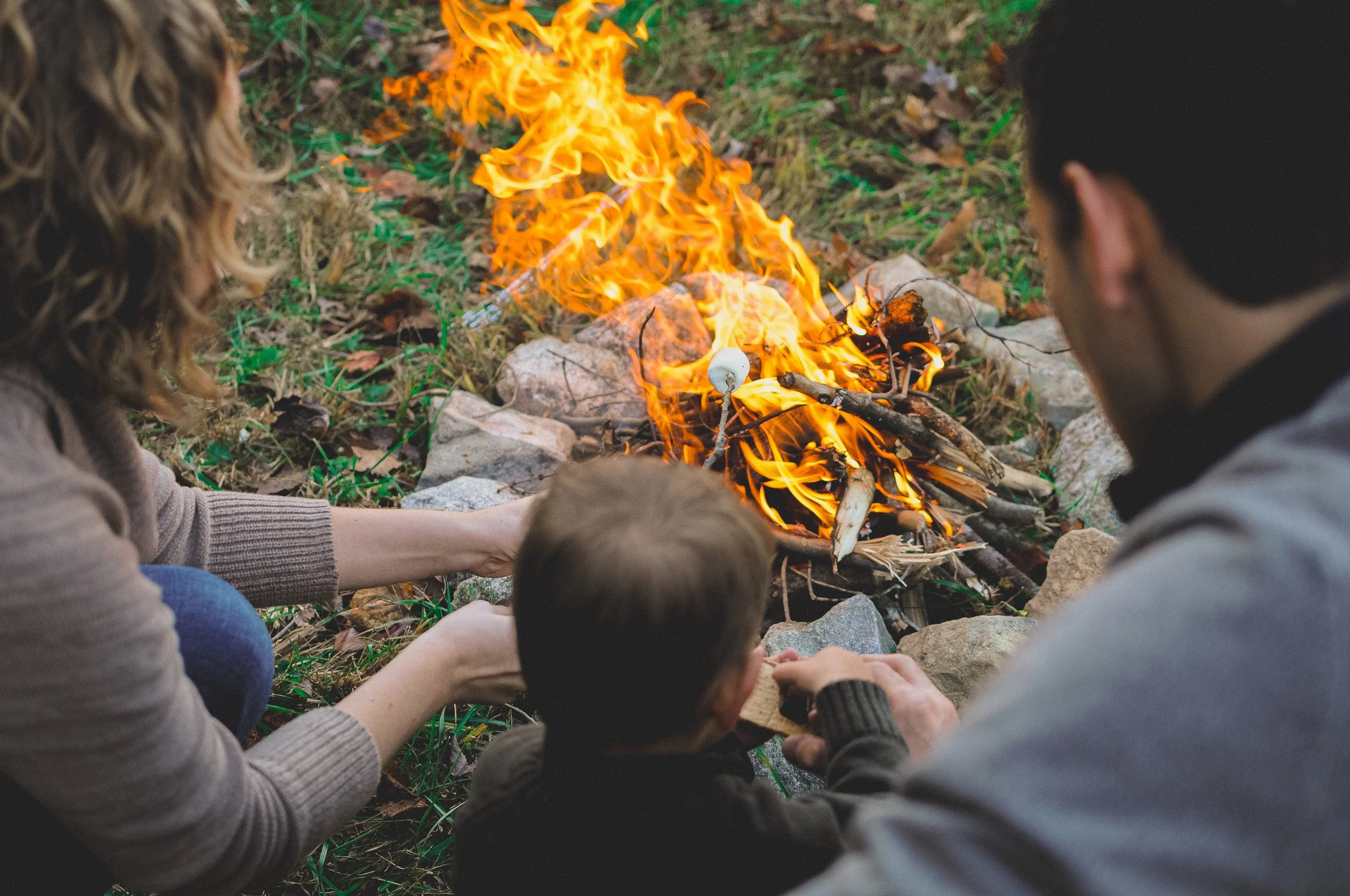 People sitting around a small fire outdoors, roasting marshmallows on sticks.