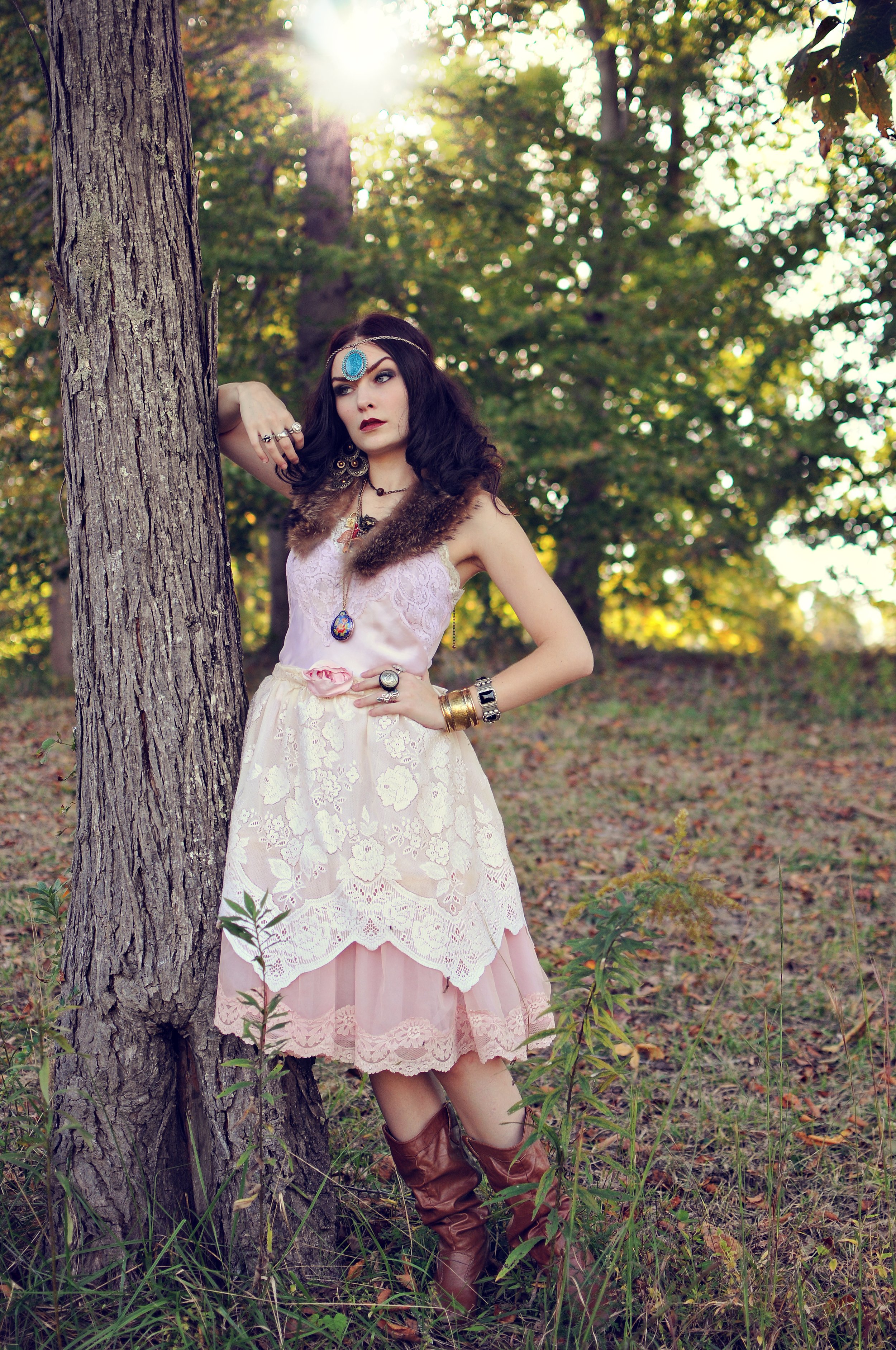 A woman wearing a bohemian style pink and cream lace dress with jewelry and brown leather boots leans against a tree in a sunlit forest clearing.