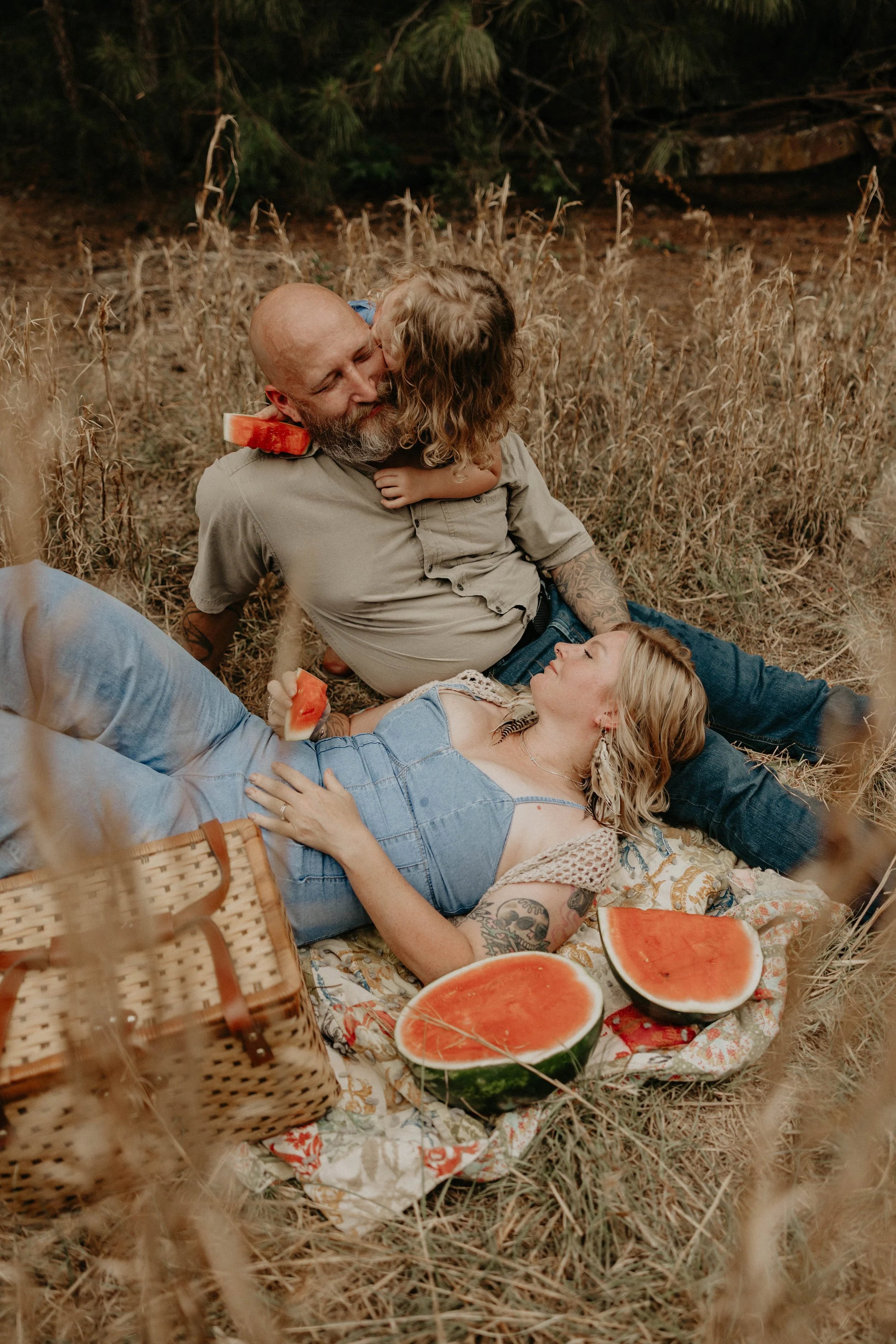 A family of four enjoying a picnic in a field of dry grass. The father, with a beard, is sitting up and embracing a young girl who is kissing him on the cheek. The mother is lying on the ground, looking up, holding a piece of watermelon. Fruit slices, including watermelons, are spread out on a blanket around them, and a woven basket is nearby.