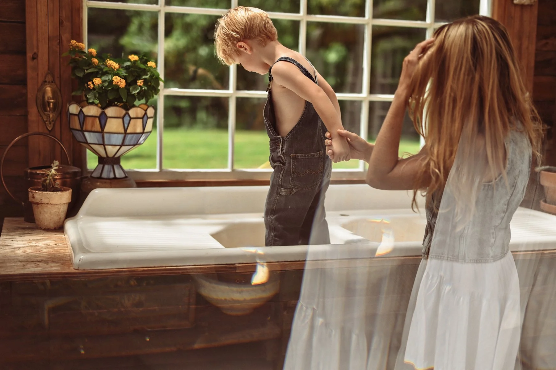 A young boy standing in a kitchen sink, with an adult woman helping him, as sunlight streams through a window. The kitchen has wooden walls, potted plants, and a marble countertop.
