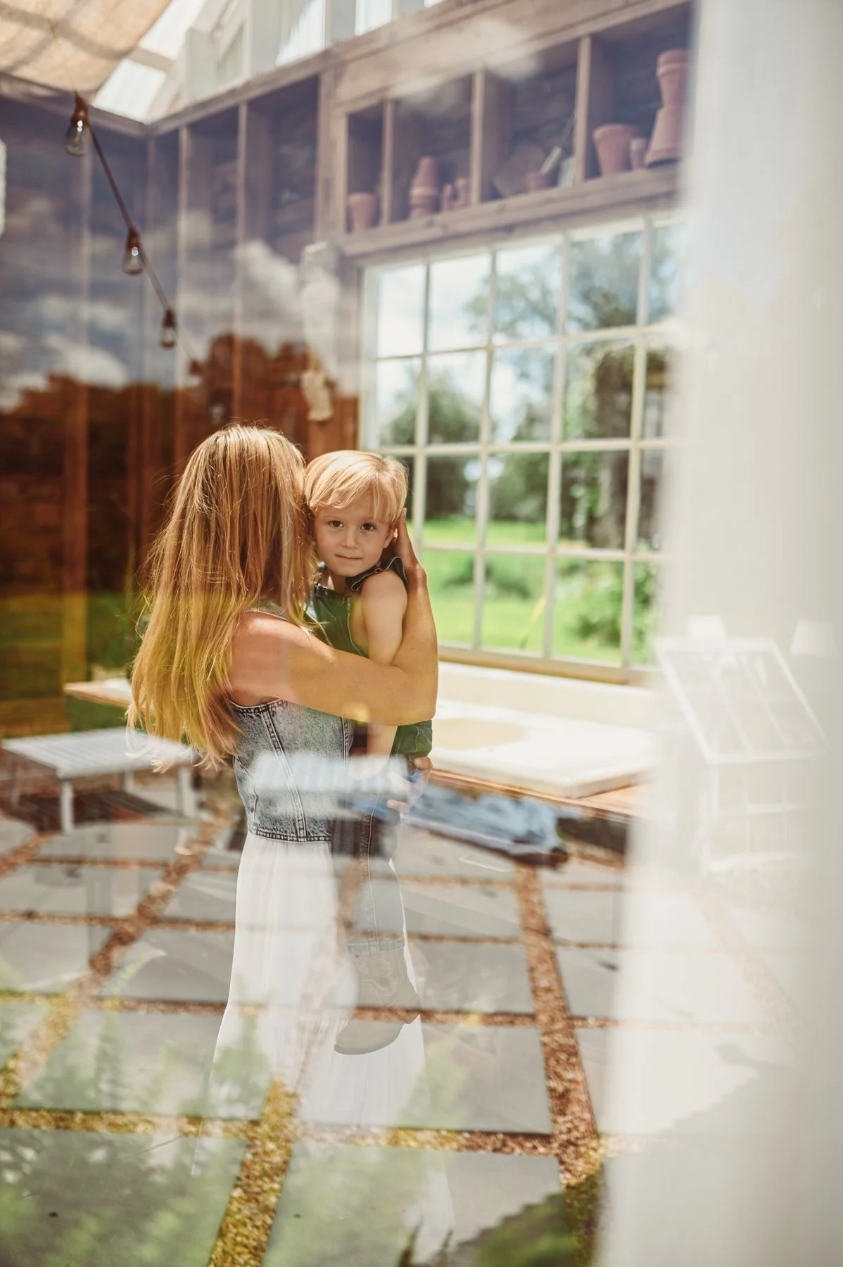 A woman with long red hair holding a young blonde boy with short hair inside a house, seen through a glass window. The house has a bright, airy feel with large window and visible outdoor greenery.