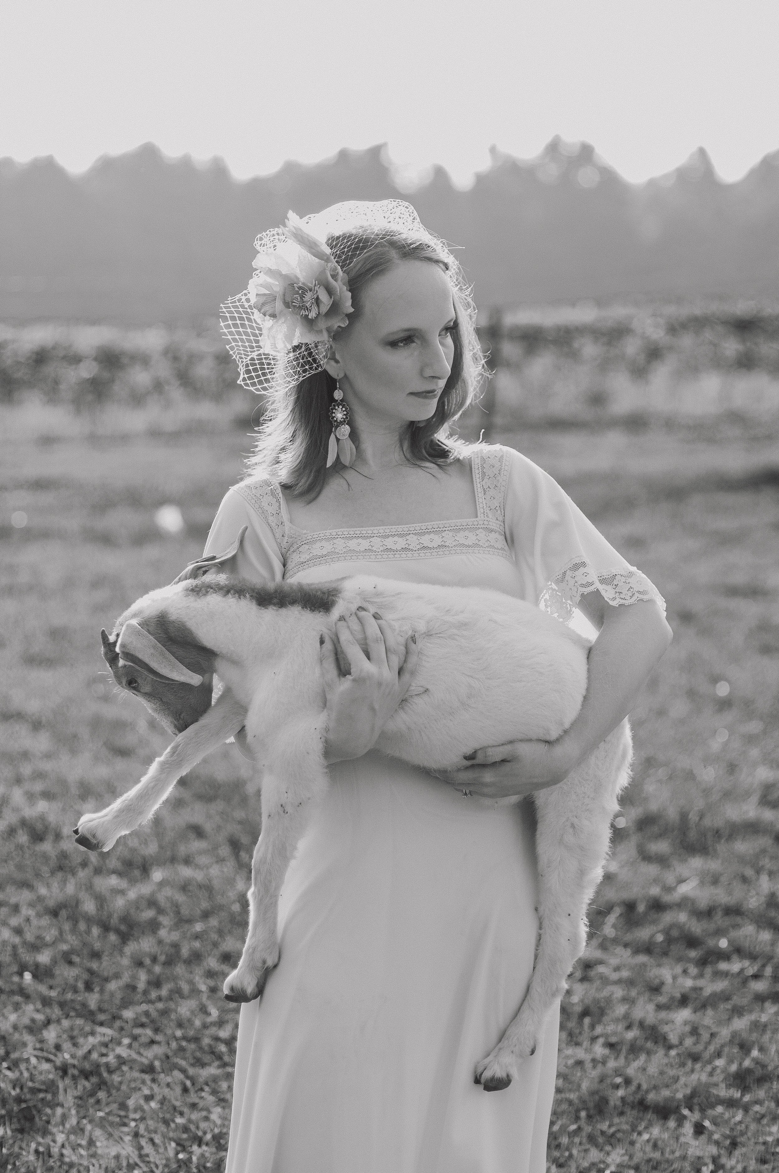 A woman in a wedding dress holding a dog in her arms, outdoors with mountains in the background, in black and white.
