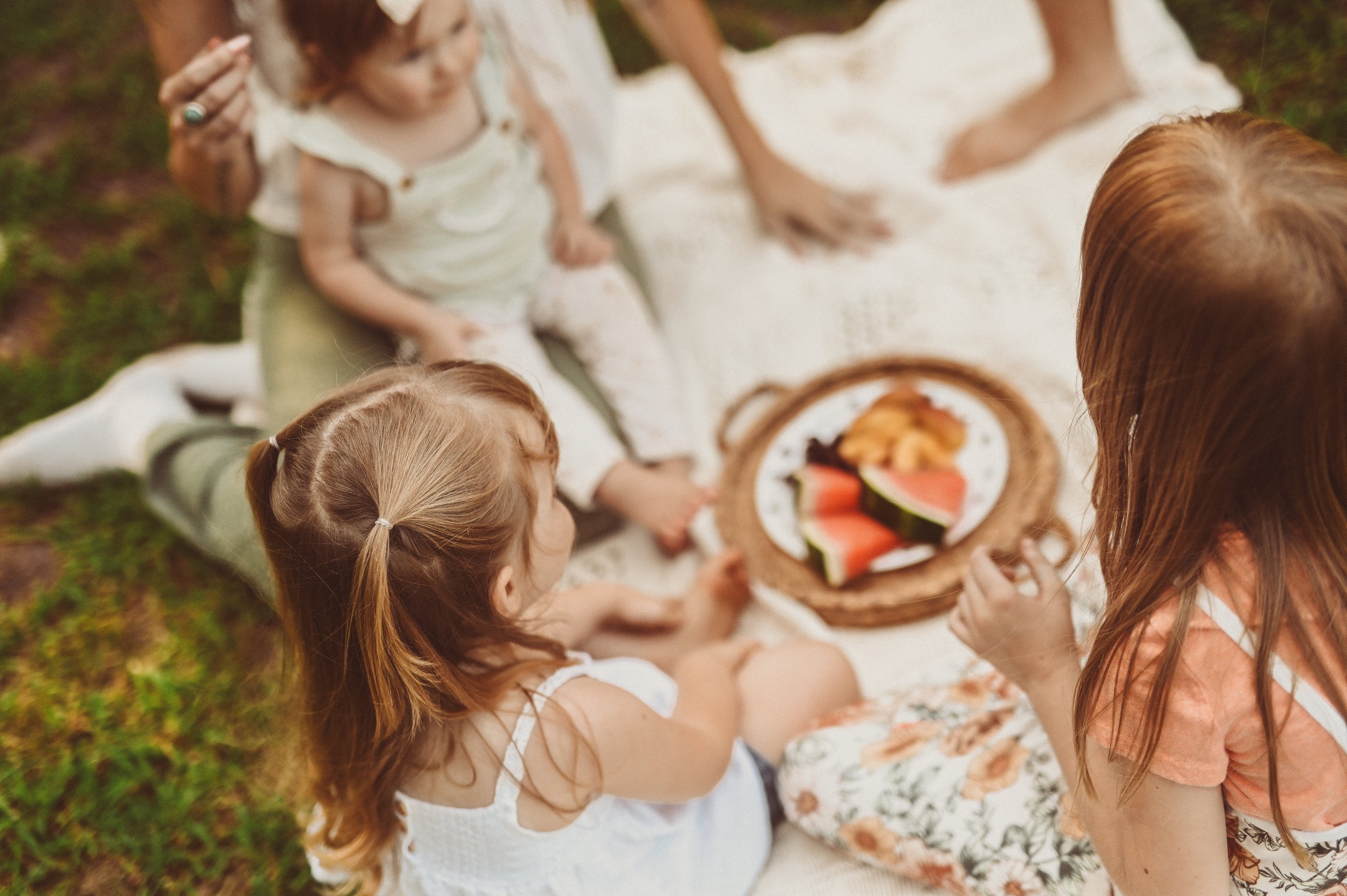 A group of children and a woman sitting on the grass, sharing a picnic with watermelon slices and other snacks.