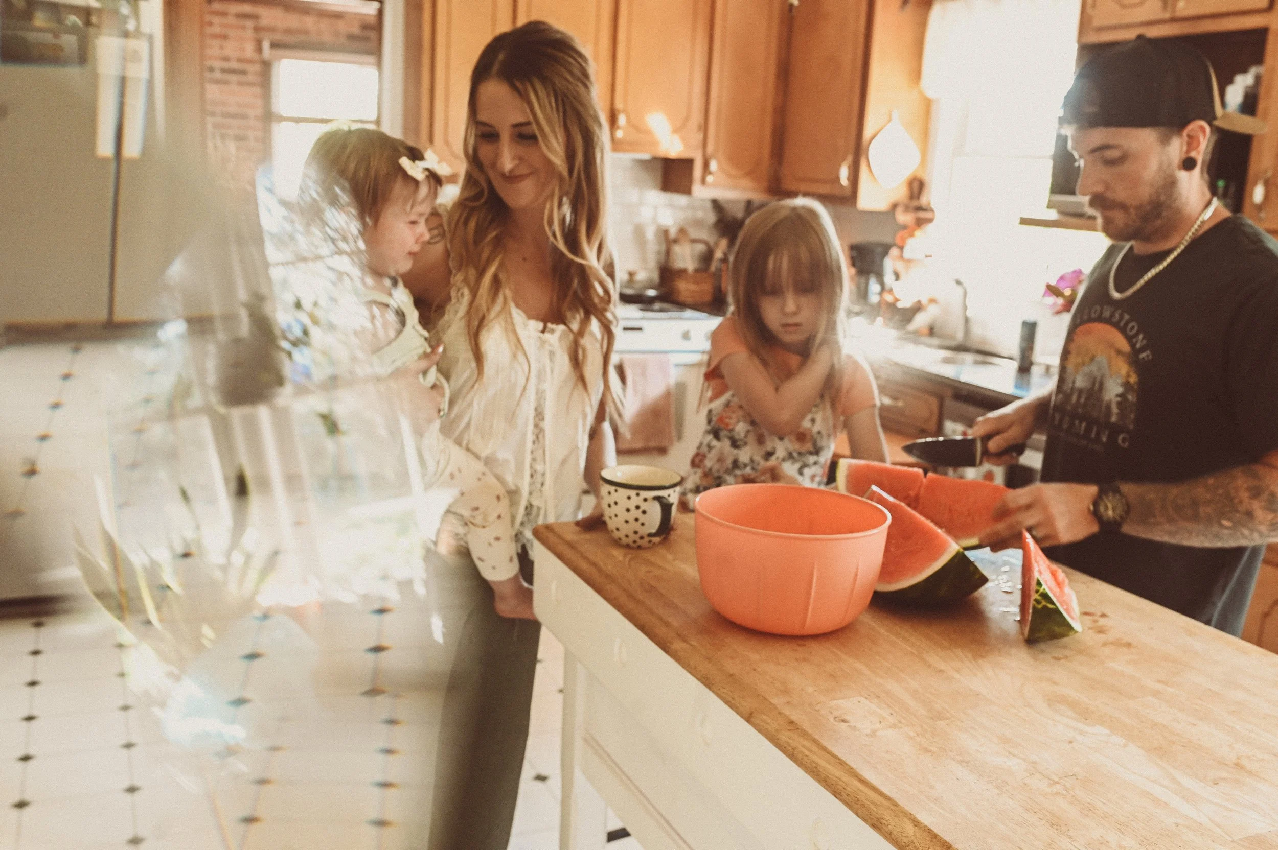 A family in a kitchen preparing sliced watermelon. A woman with long wavy hair is holding a young girl with floral clothing, while a man with a cap and earrings uses a knife to cut the watermelon. The kitchen has wooden cabinets and a window letting 