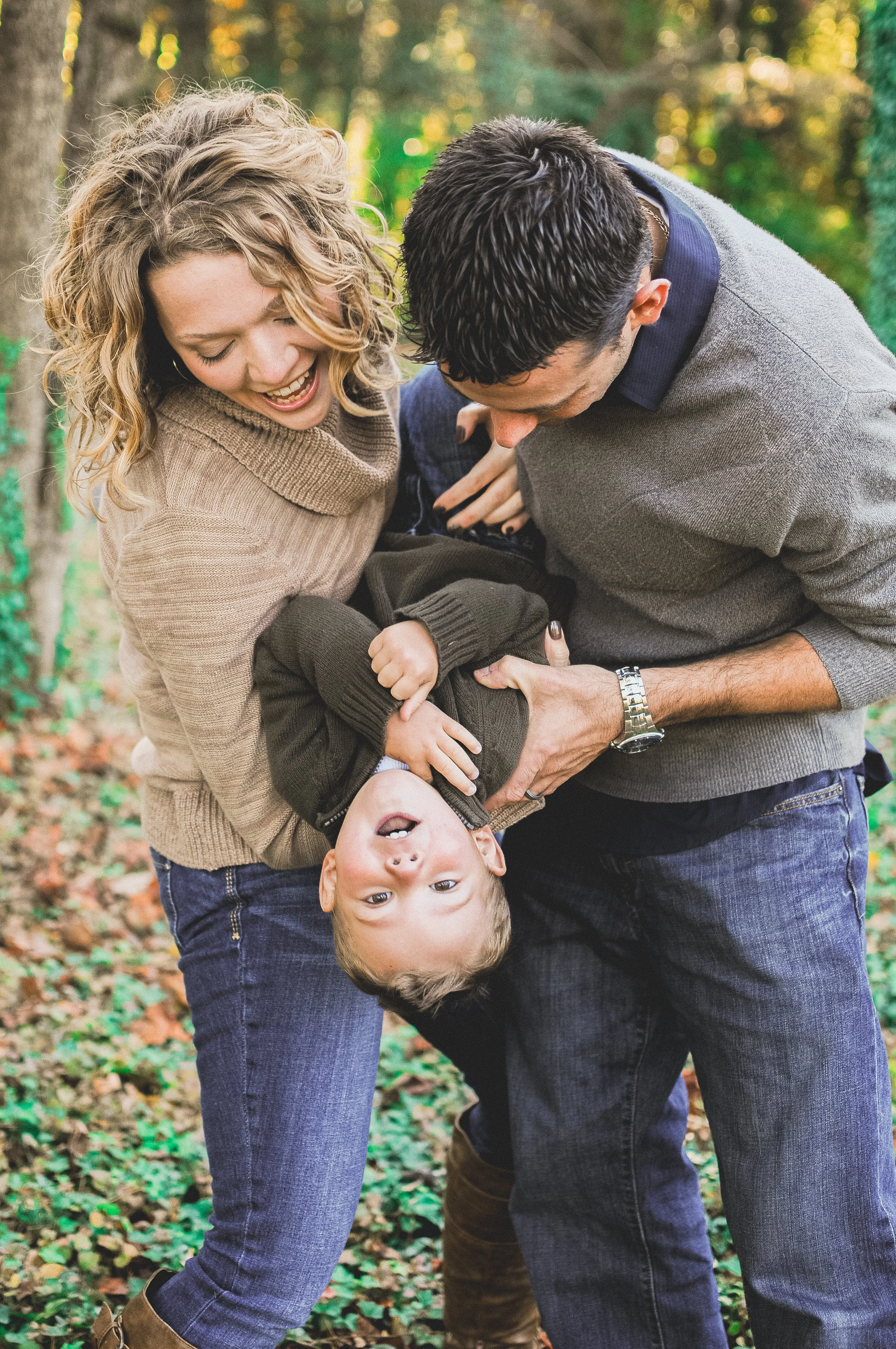 Family of three, two adults and a young boy, playfully leaning backwards outdoors in a forested area with fallen leaves on the ground, smiling and laughing.