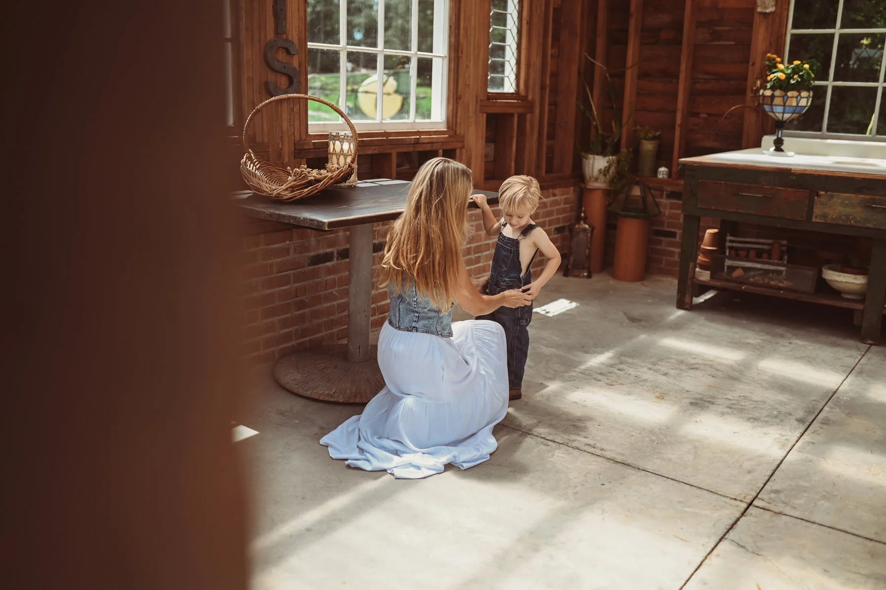 A woman kneeling on the ground helping a young boy put on his overalls inside a rustic sunlit barn with wooden walls and large windows.