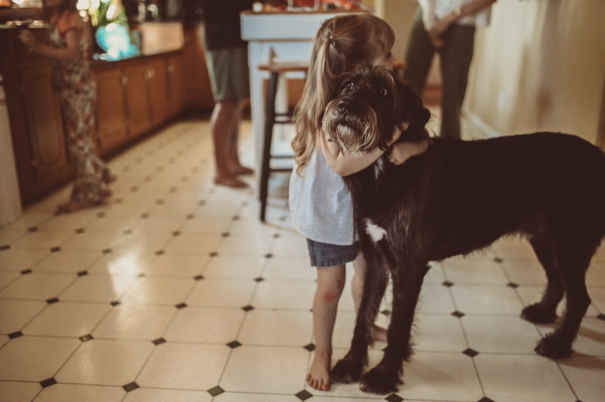 A young girl hugging a large black and white dog, standing barefoot on a tiled kitchen floor.