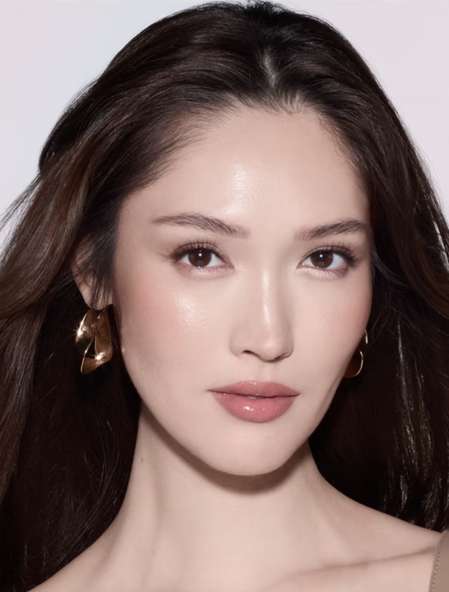 Close-up of a woman with long, dark brown hair, light makeup, and gold hoop earrings, looking directly at the camera.
