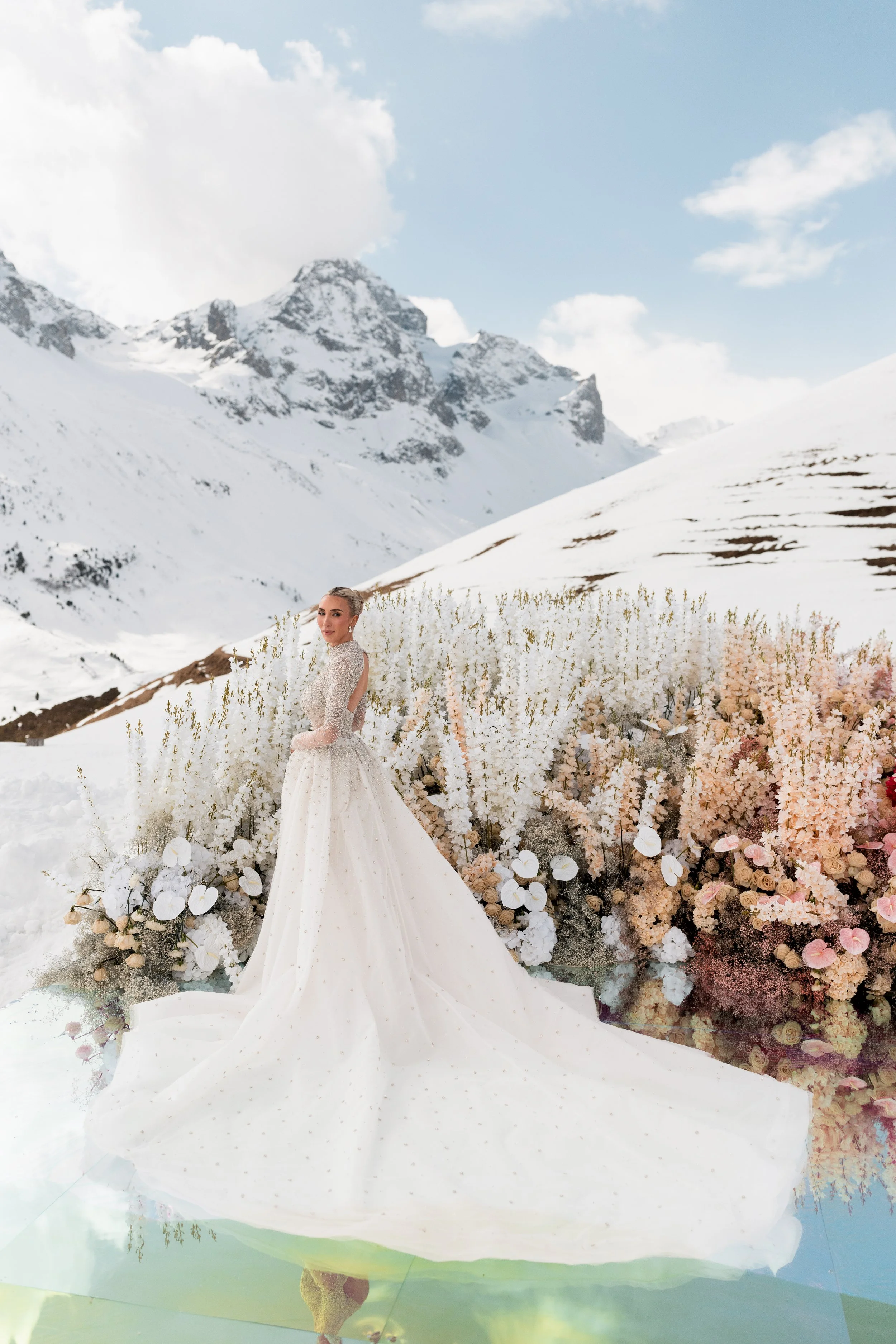 A bride in a white wedding gown standing in front of snow-covered mountains and a flower display with pink, white, and peach blossoms.