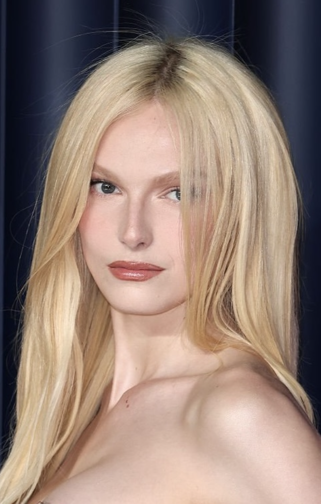 A portrait of a woman with long blonde hair, light skin, and a subtle smile, set against a dark curtain background.