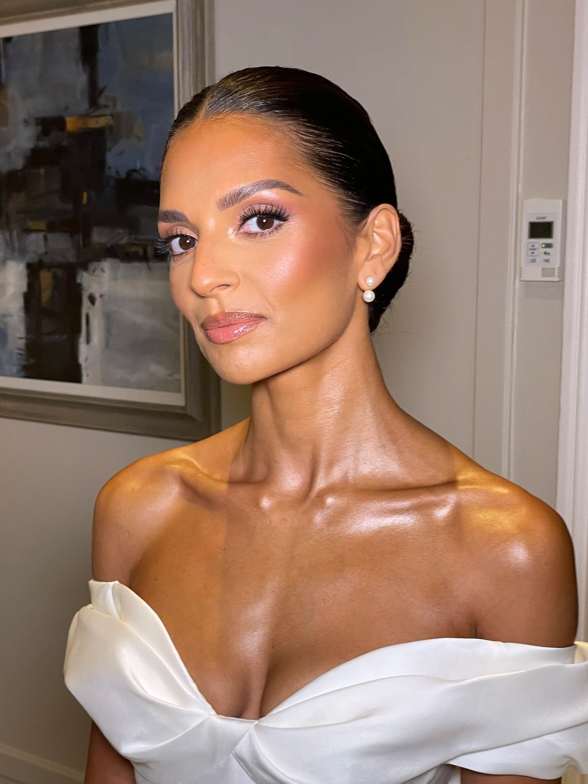 A woman with smooth, dark hair styled in a sleek bun, wearing white pearl earrings. She has makeup with well-defined eyebrows, long eyelashes, and a glossy nude lipstick. She is dressed in a white off-shoulder gown.