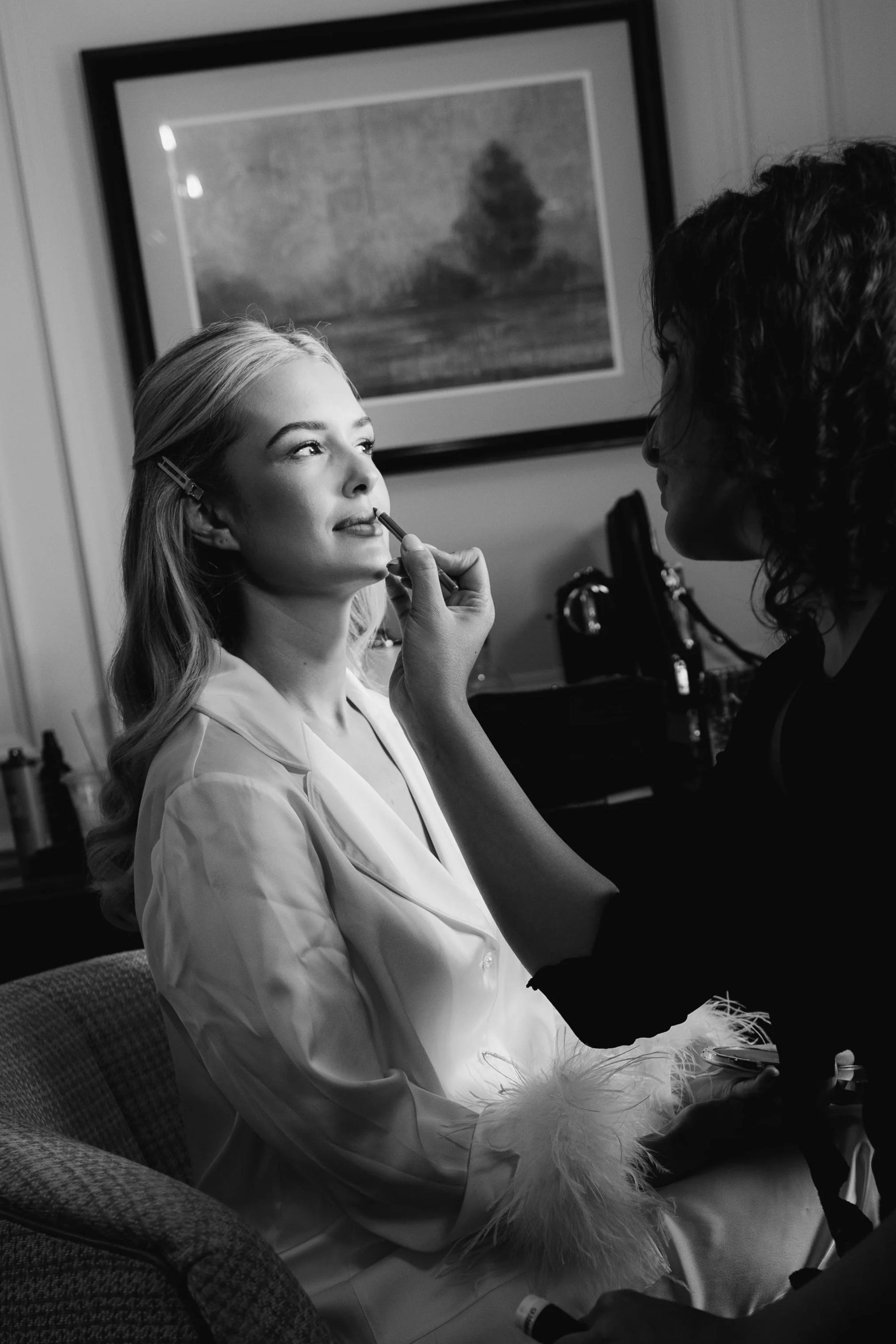 A woman with blonde hair, wearing a white jacket, sitting on a chair while a makeup artist applies lipstick to her lips. The scene appears to be in a hospital or clinical setting, with a framed painting of a tree and landscape in the background.
