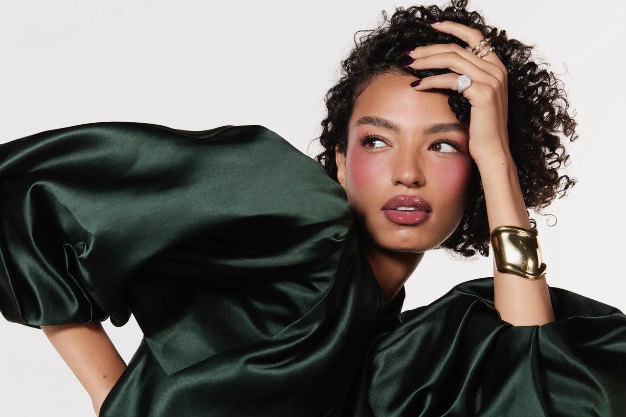 A woman with curly hair wearing a dark green satin dress, gold bracelet, and rings, posing with her hand on her forehead against a plain white background.