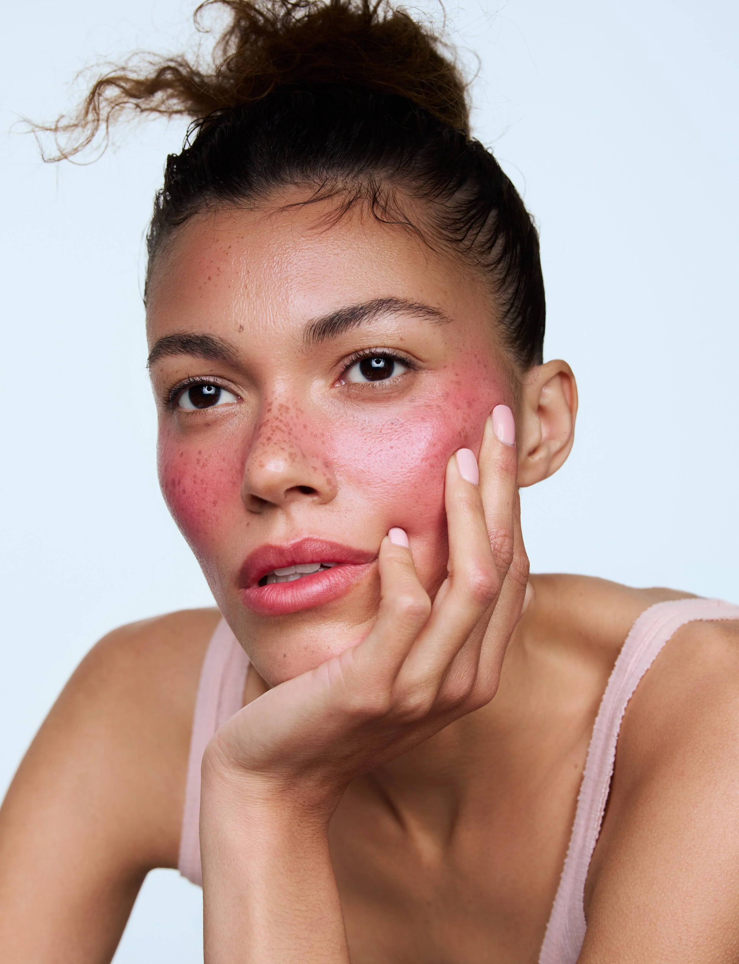 Close-up of a young woman with natural makeup, freckles, and red cheeks, wearing a light pink tank top, with her hand gently touching her face, against a plain light blue background.